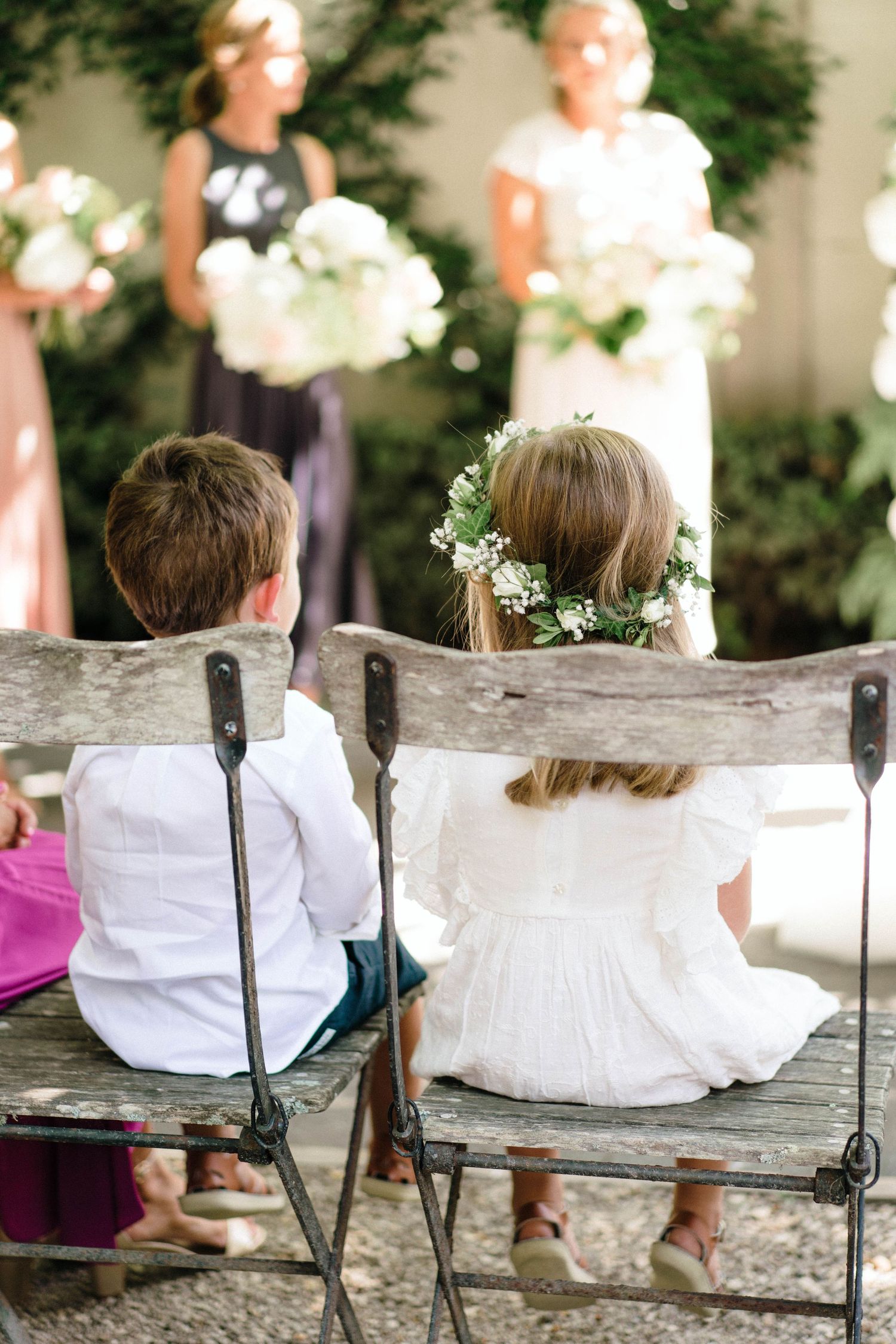 Children in white dresses sit together on a rustic wooden bench in a garden setting during a special occasion.