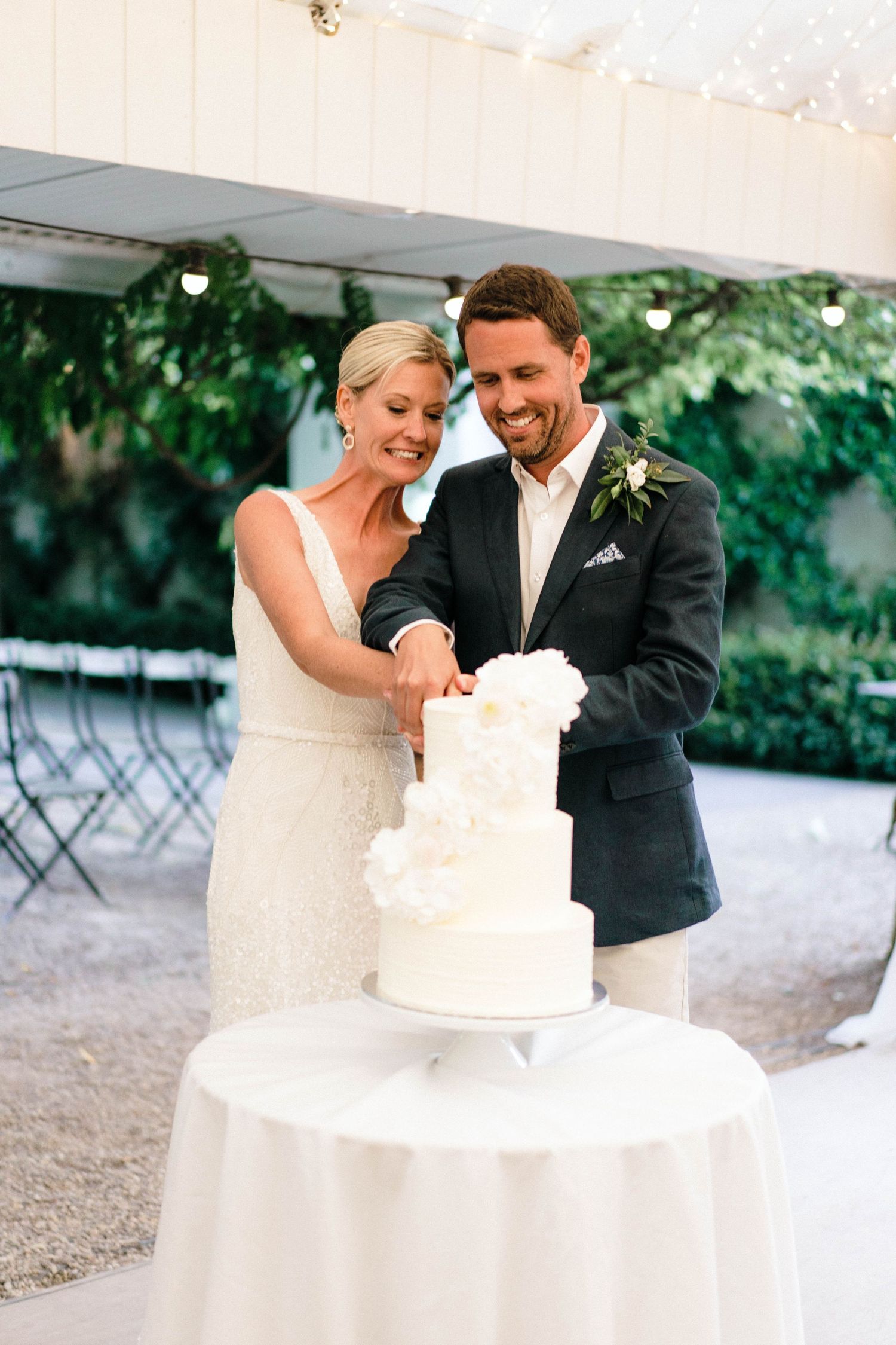 Couple shares sweet moment while cutting their elegant white wedding cake outdoors.