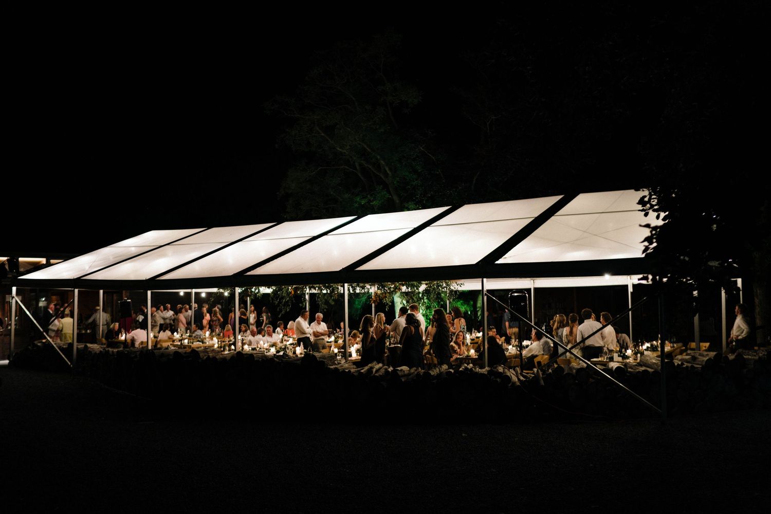 An illuminated outdoor wedding reception tent glows at night with guests dining inside.