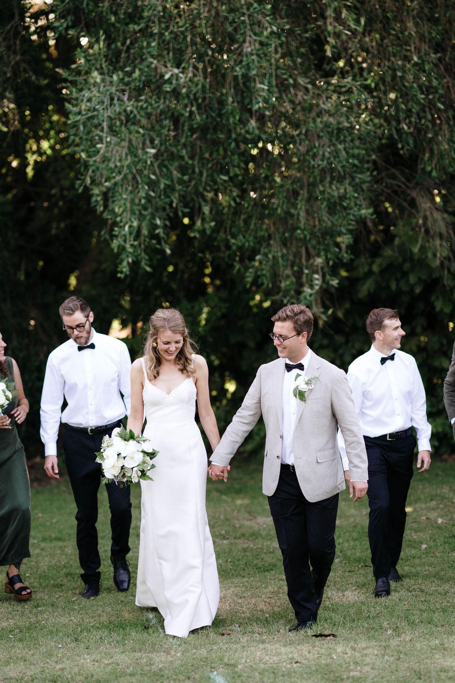 Wedding party walks together on grass with trees in background during outdoor celebration.