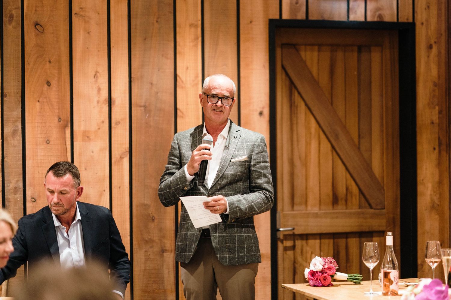 A speaker in a plaid blazer addresses guests during a dinner event at a rustic wooden venue.