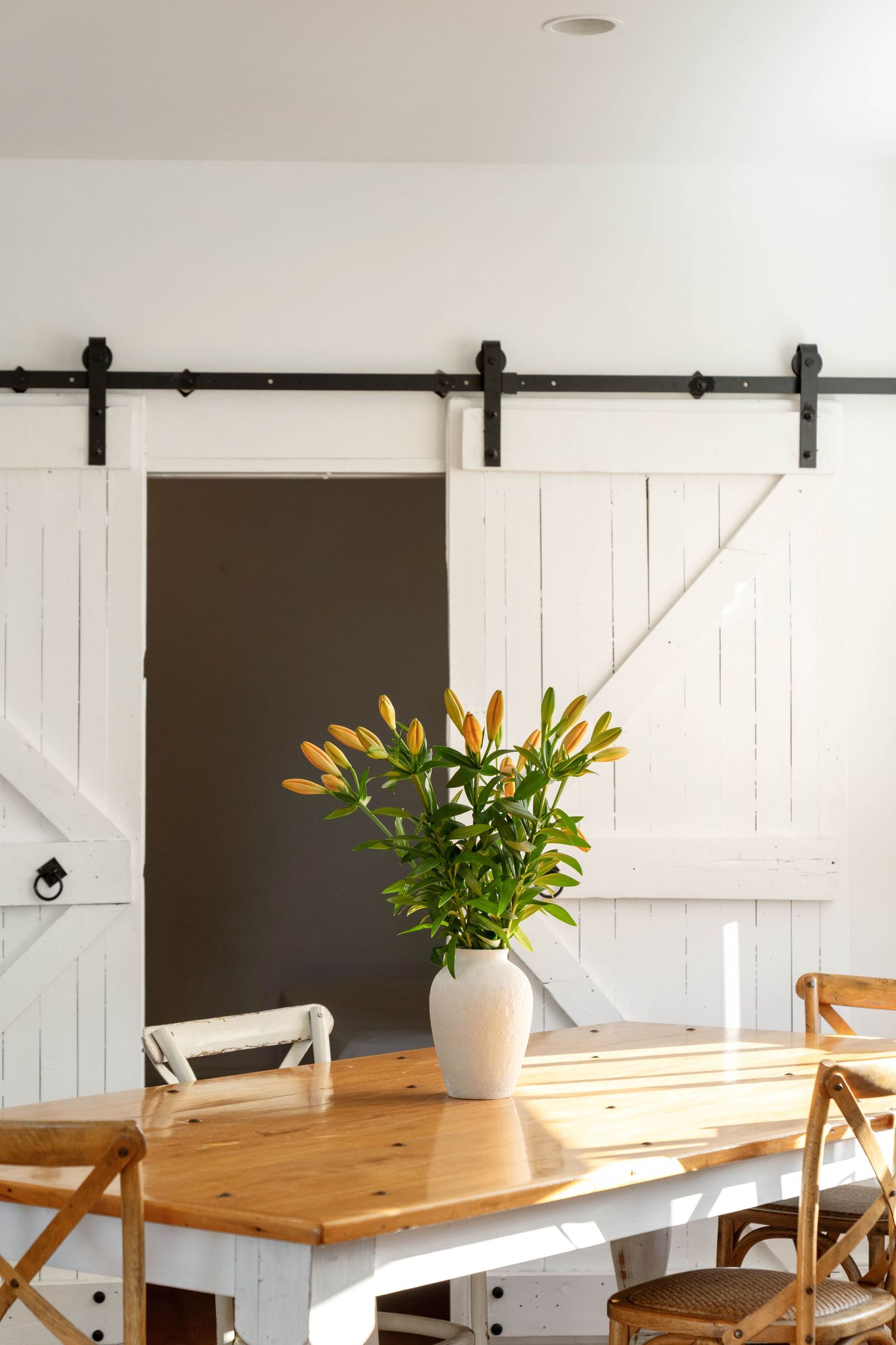 White barn doors with sliding hardware frame a dining area featuring yellow lilies in a vase on a wooden table.