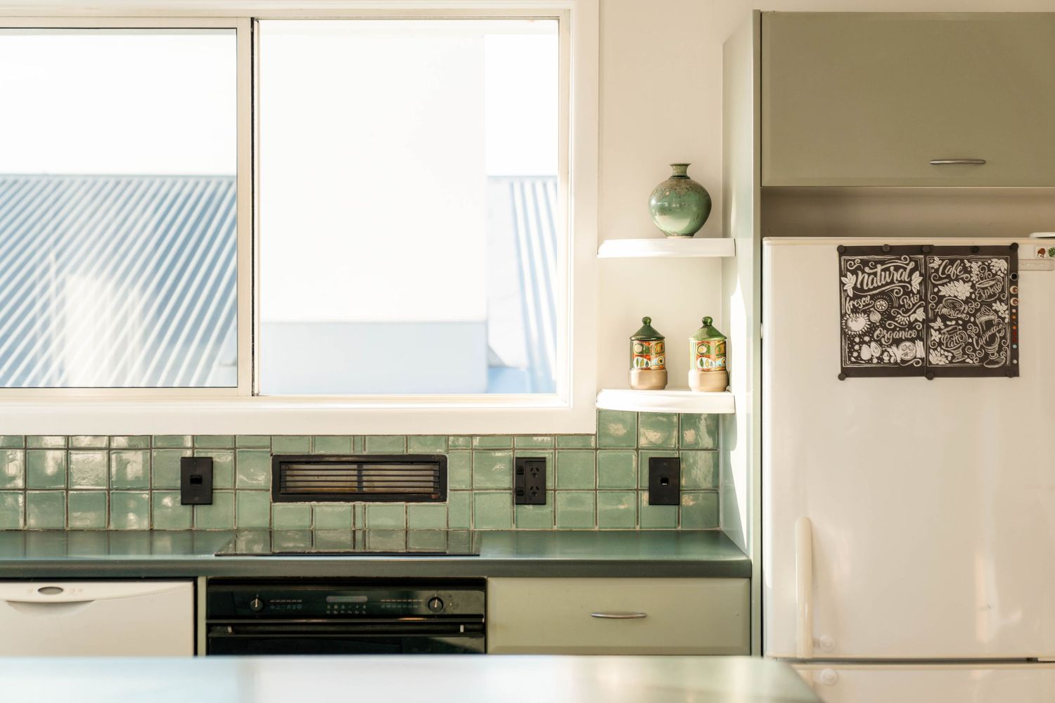 A minimalist kitchen with mint green tile backsplash features a window, white cabinets, and hanging storage.