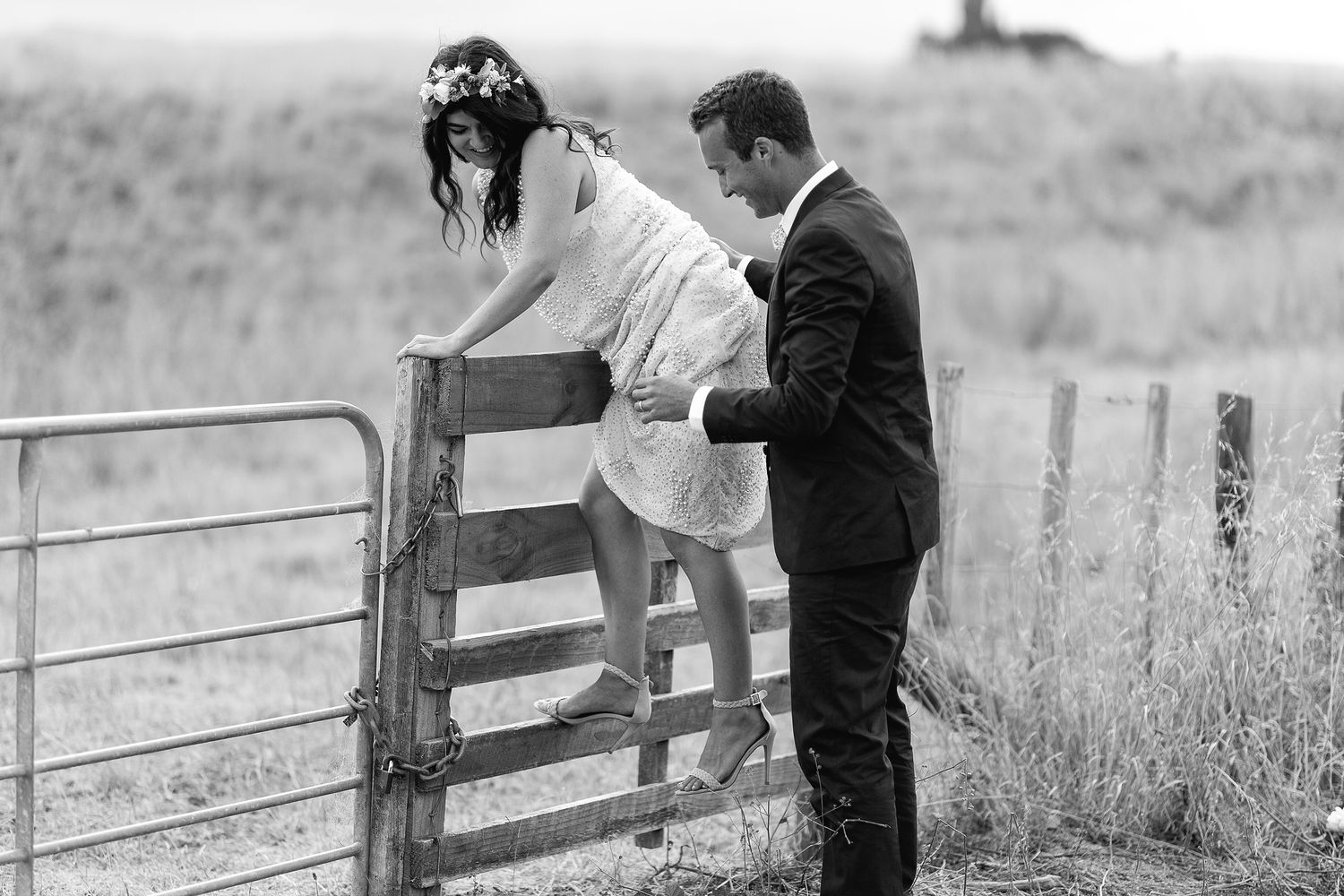 A couple shares a romantic moment together in a monochrome rural setting with wooden fences and tall grass.