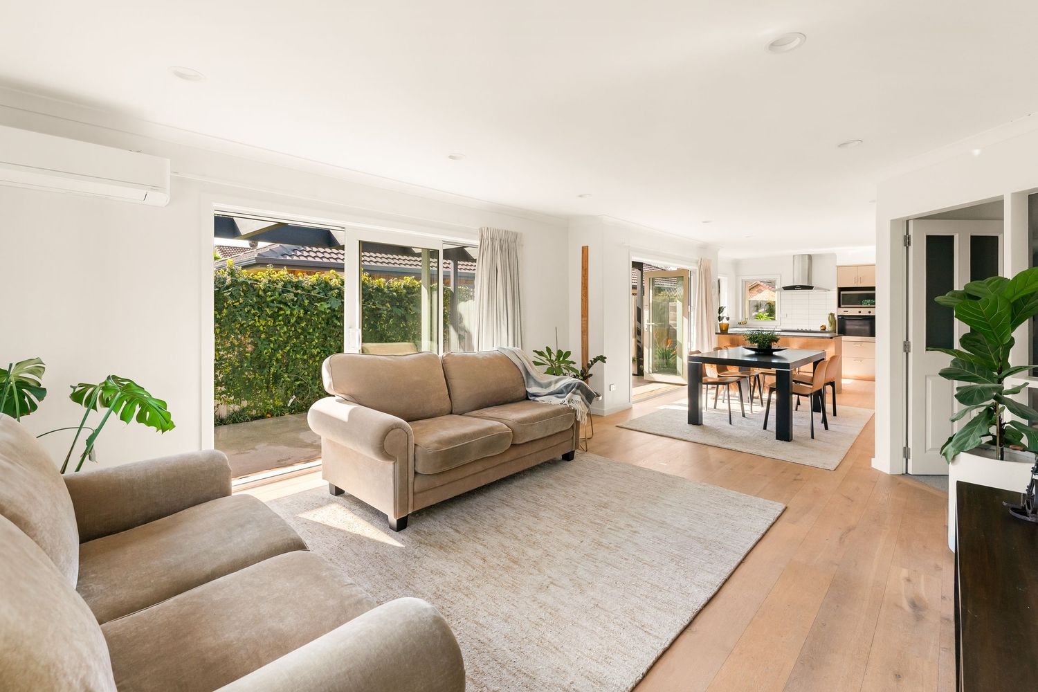 Modern open concept living room with beige sofas, hardwood floors and large windows overlooking green hedges.