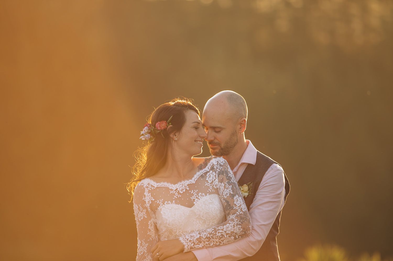 A romantic couple embraces during golden hour sunset, with the bride wearing a lace dress and groom in a white shirt and vest.