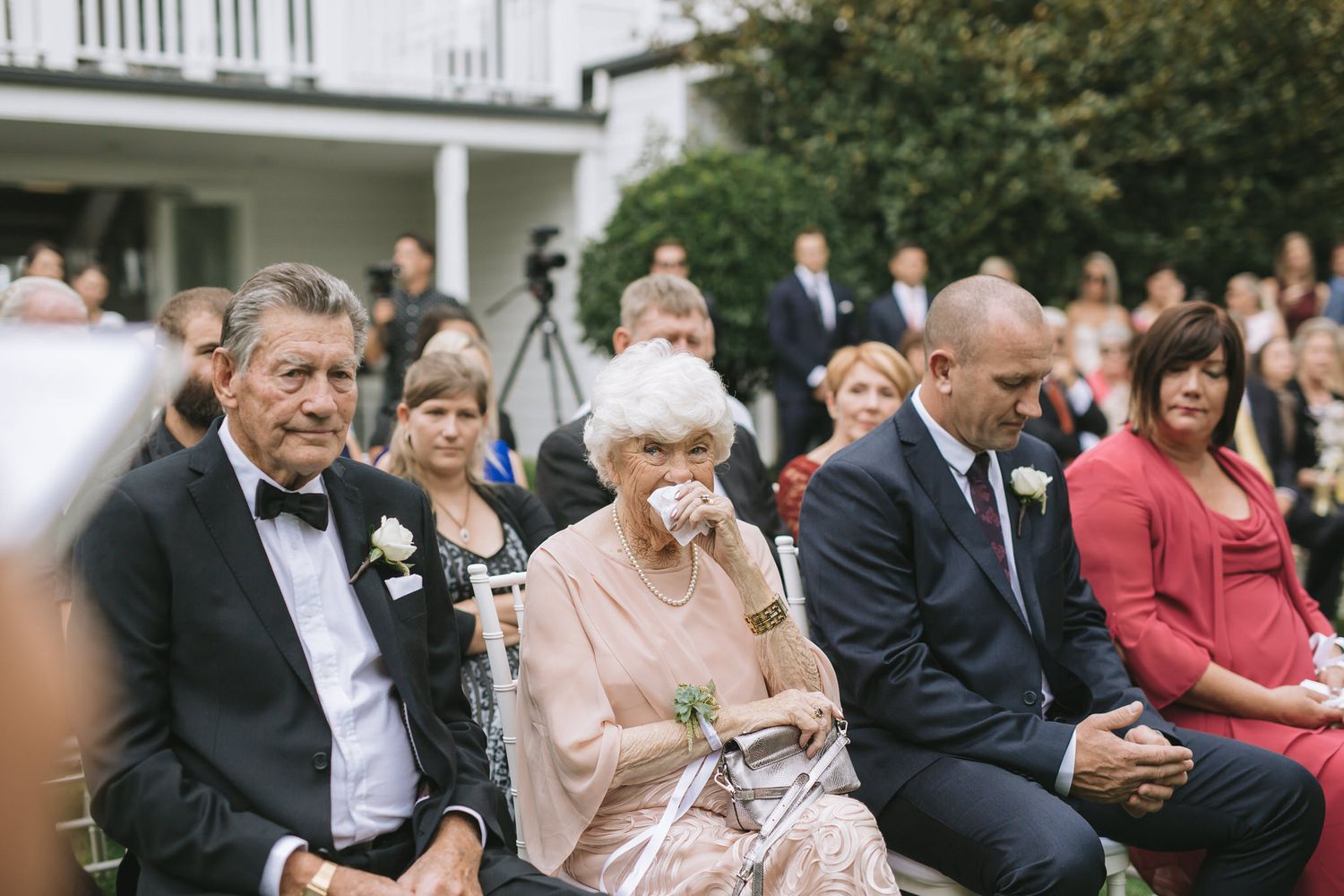 Wedding guests seated in rows outdoors for a ceremony with formal attire and flowers on a sunny day.