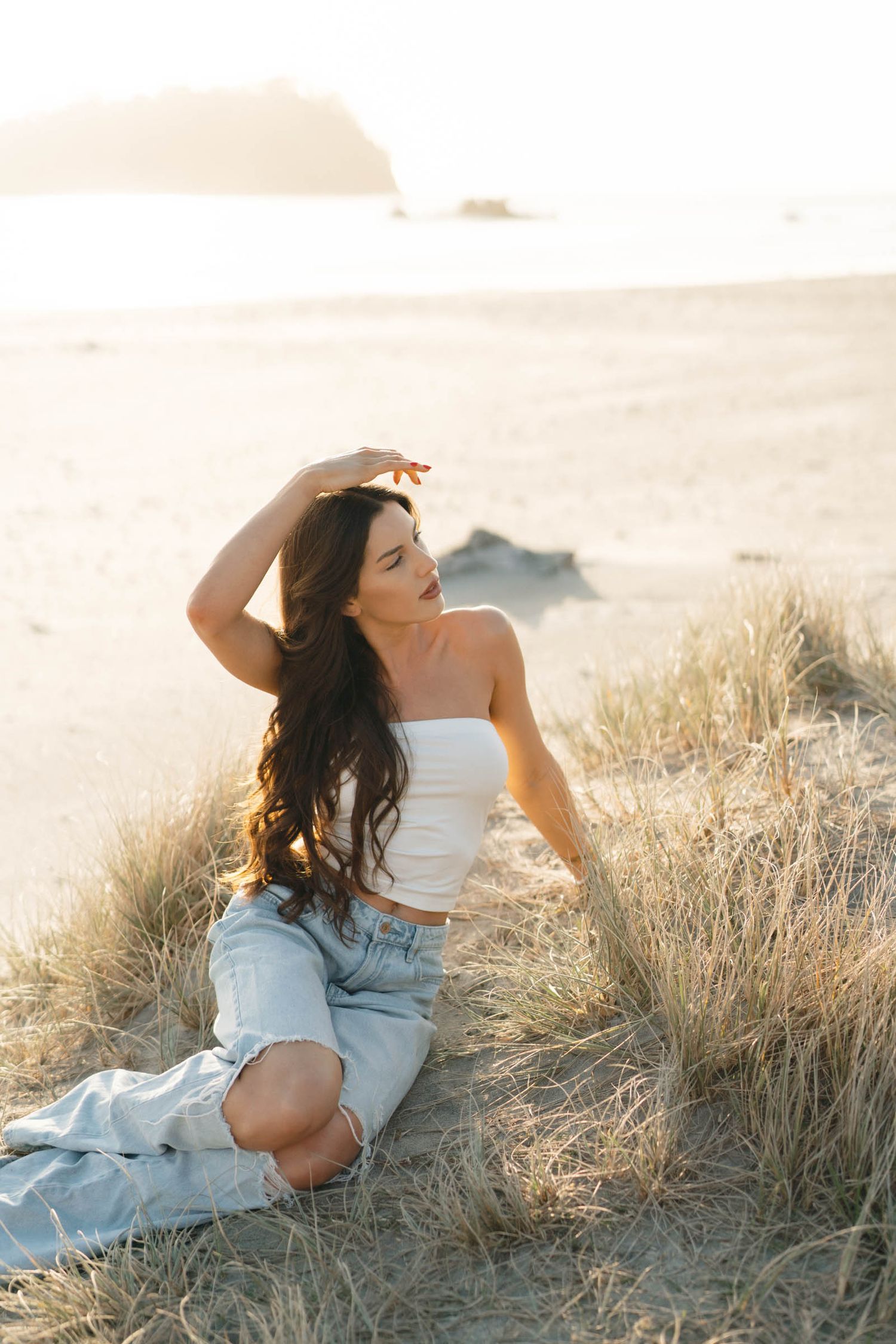 Person in white top and denim sits among beach grass during sunset.
