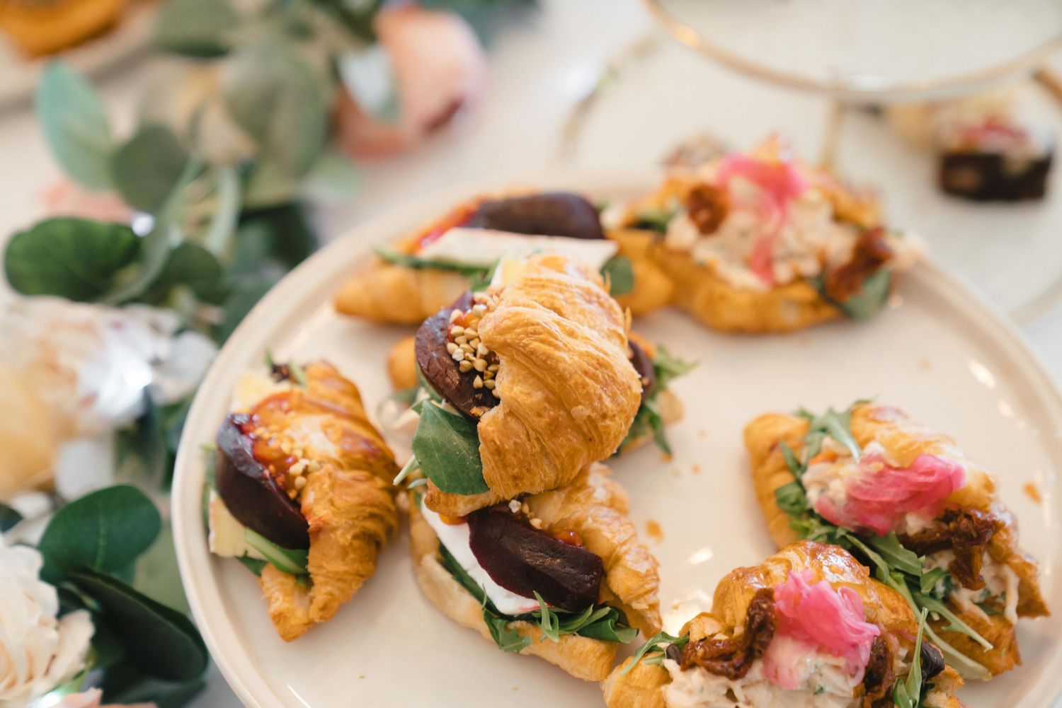 Gourmet mini croissant sandwiches with fresh greens and edible flowers served on a light beige plate.