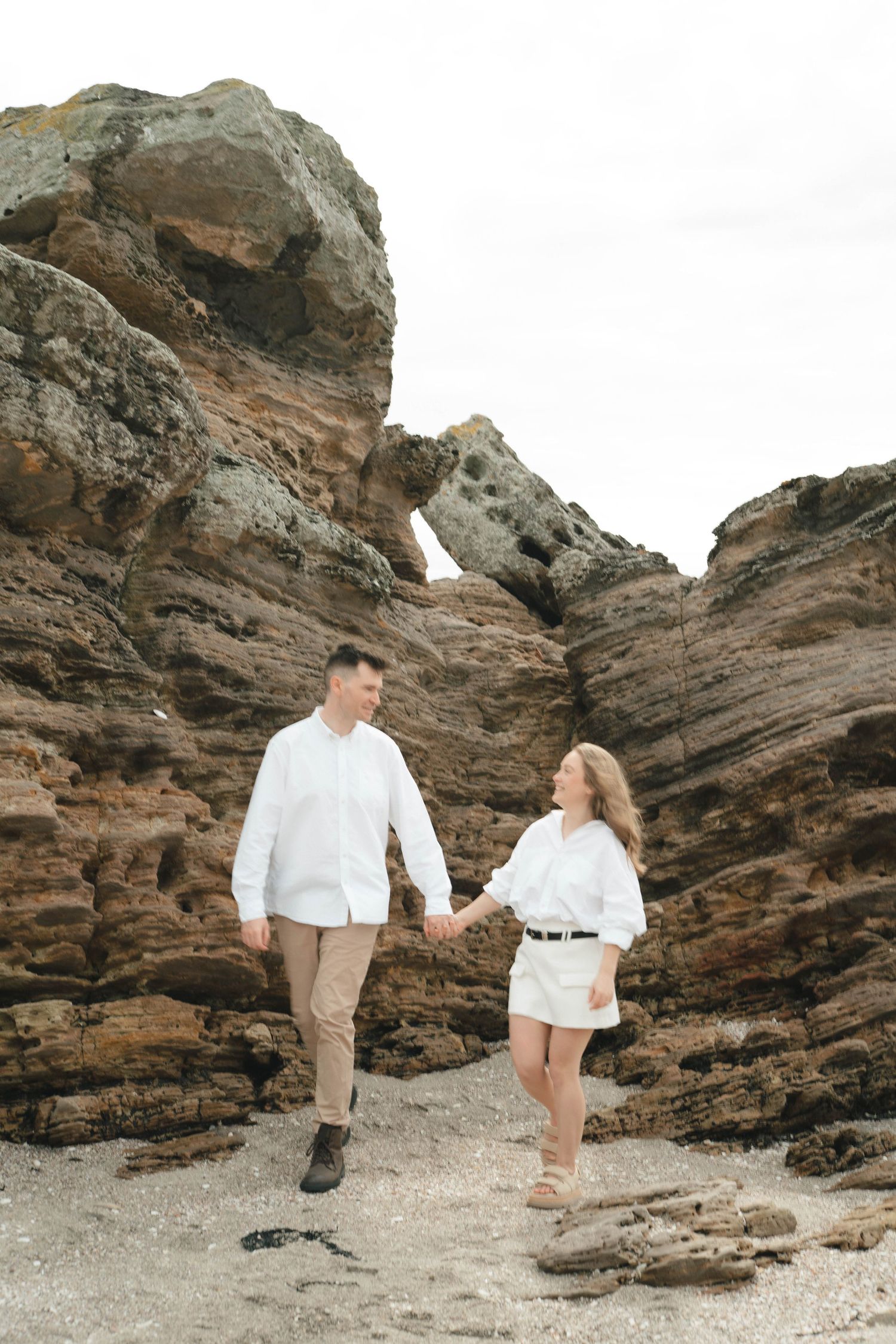Two people in white outfits walk hand in hand along a dramatic rocky beach landscape.