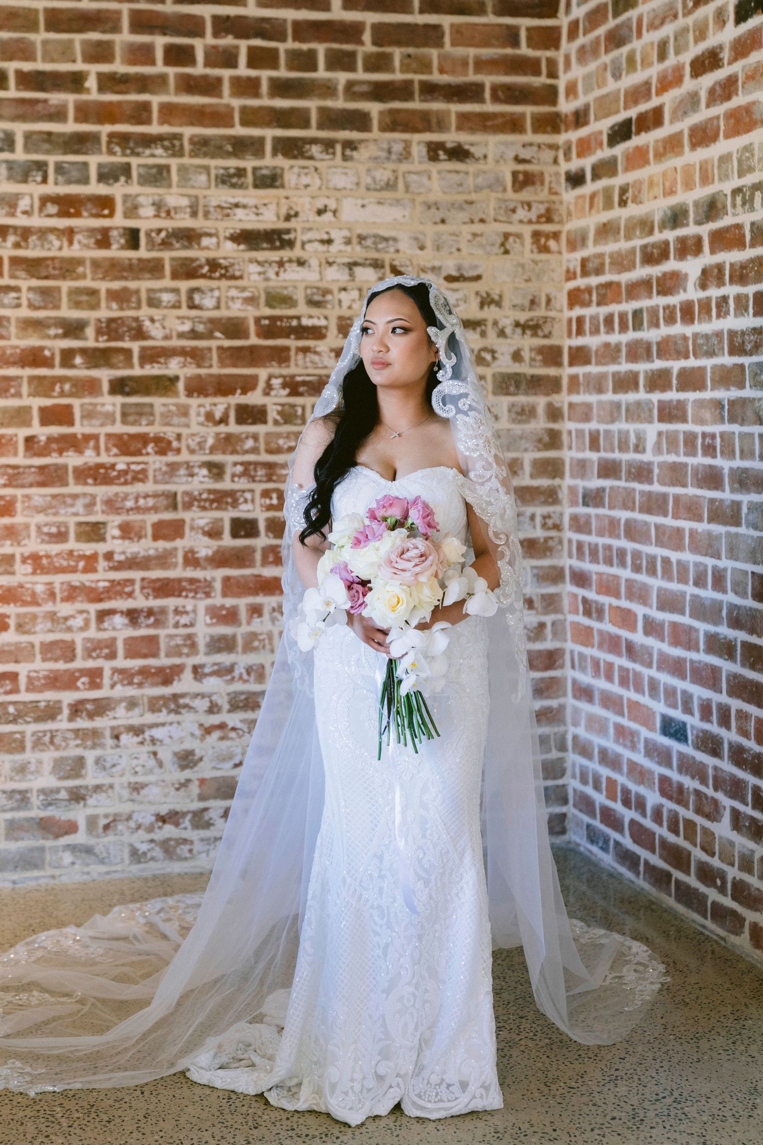 A stunning bride in a lace wedding gown holds a pink and white bouquet against a rustic brick wall.