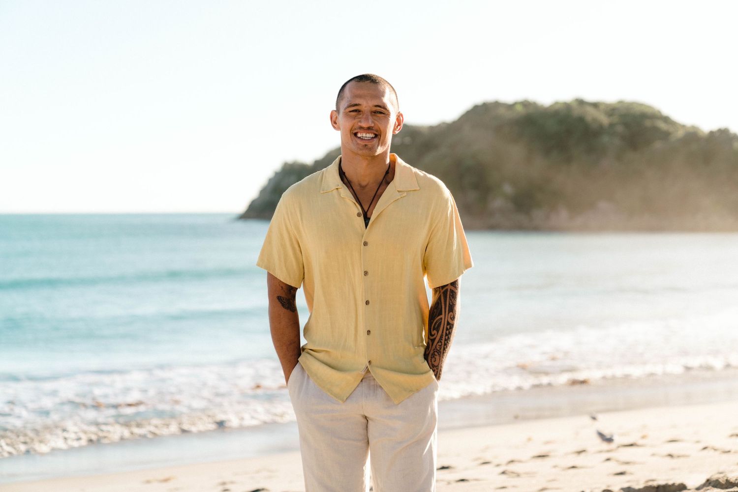 A person wearing a yellow button-up shirt and light pants stands smiling on a sunny beach with ocean and cliffs behind.
