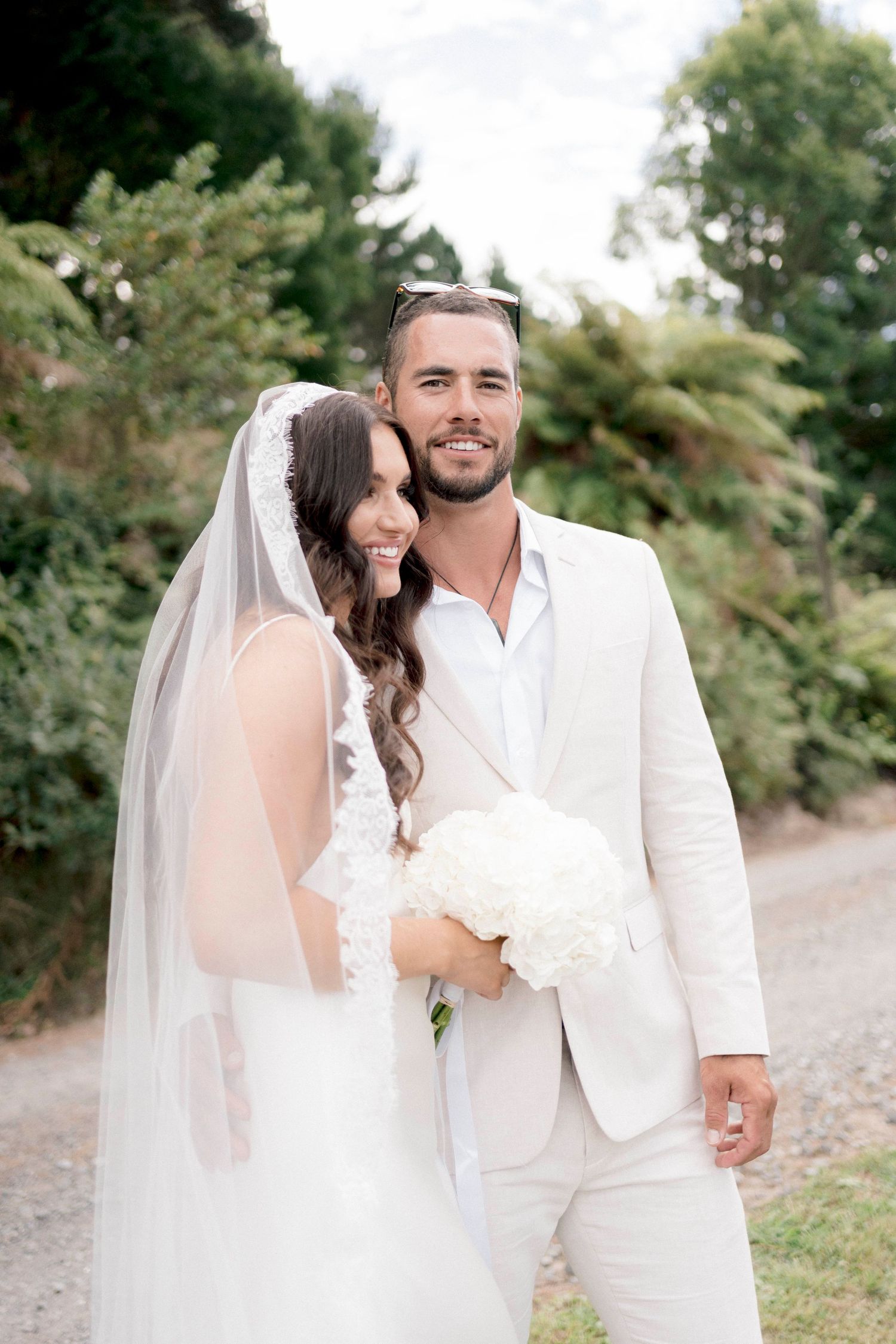 Newlyweds share an intimate moment in a garden setting, both dressed in elegant white attire.