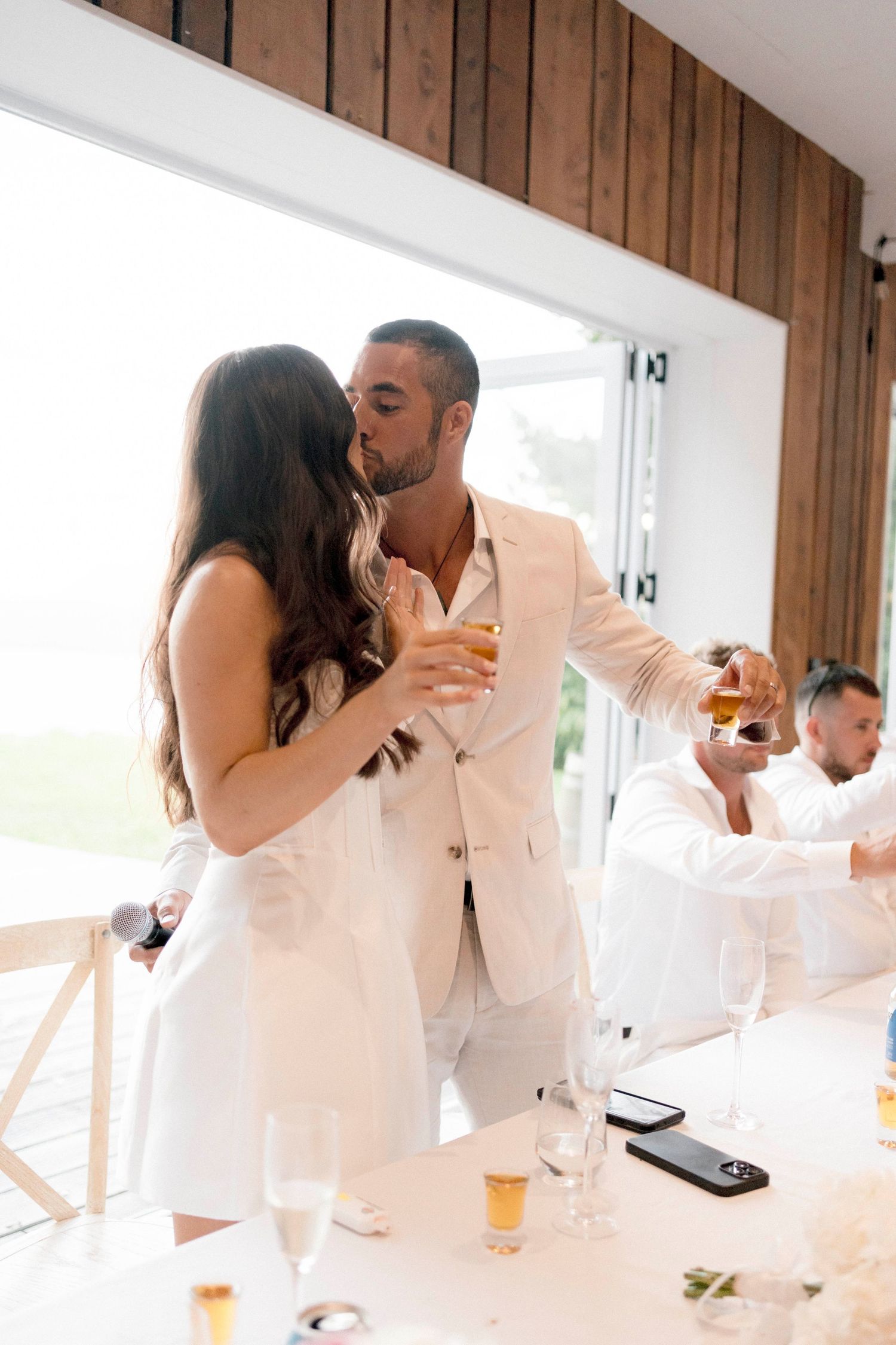 A romantic moment between a couple in white attire sharing drinks at an elegant reception.