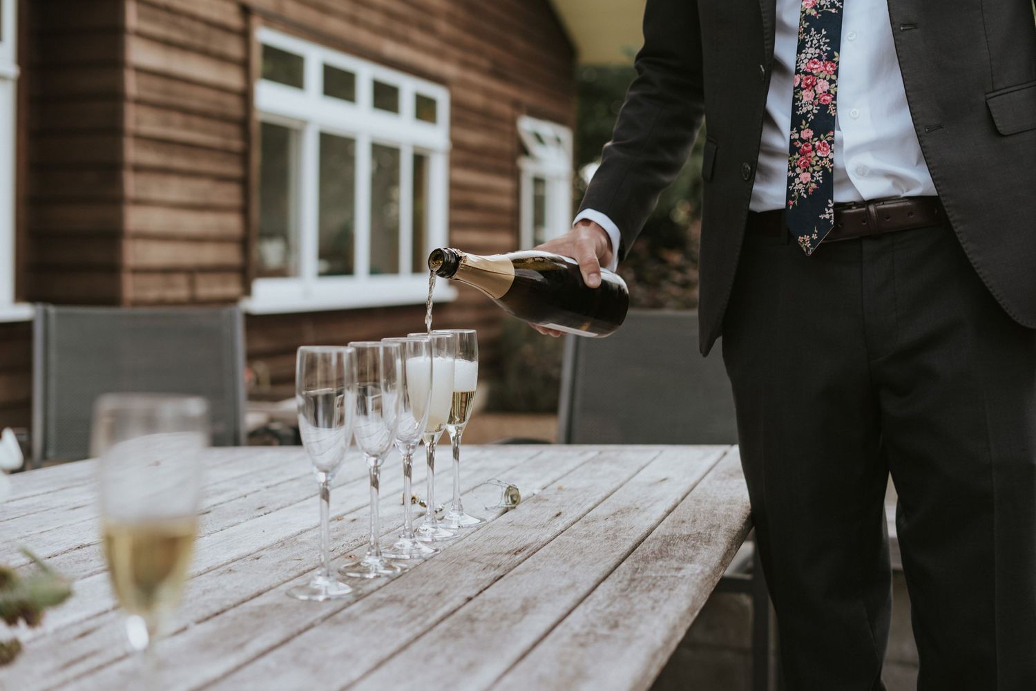 Someone pours champagne into elegant glass flutes arranged on a wooden table during an outdoor celebration.