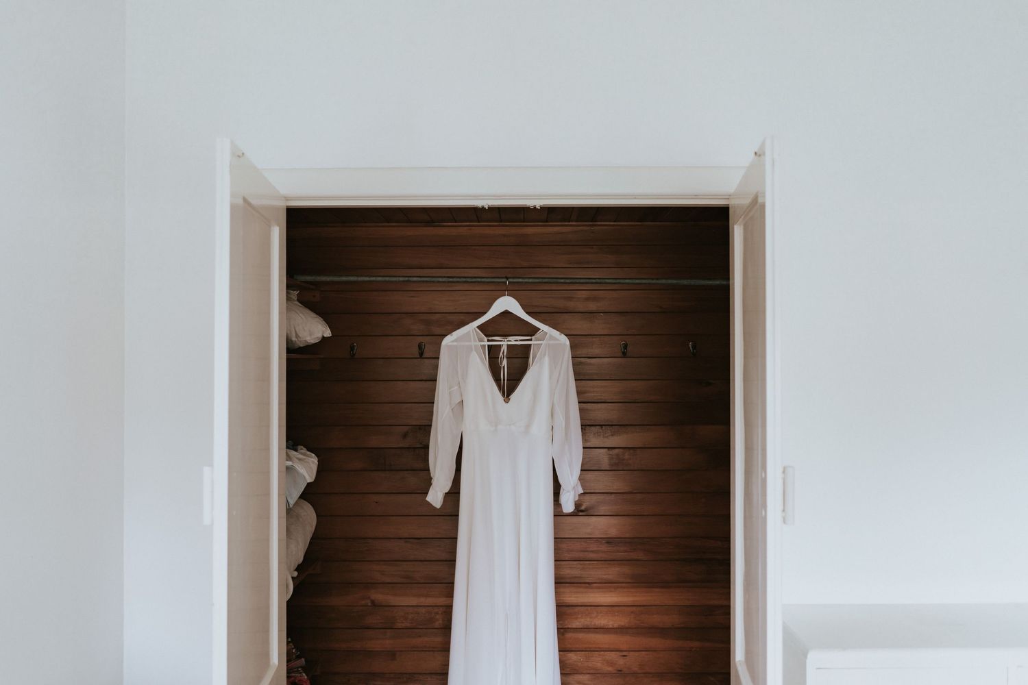 Simple white wedding dress hanging on wooden wall in natural lighting.