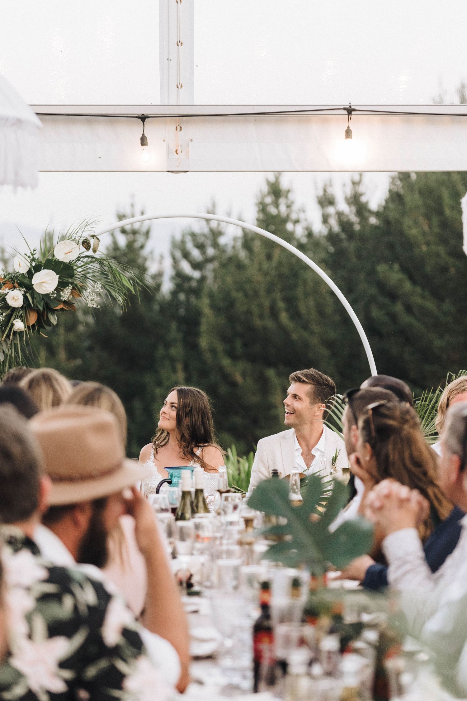 Wedding reception dinner set under a clear tent with string lights and floral decorations.