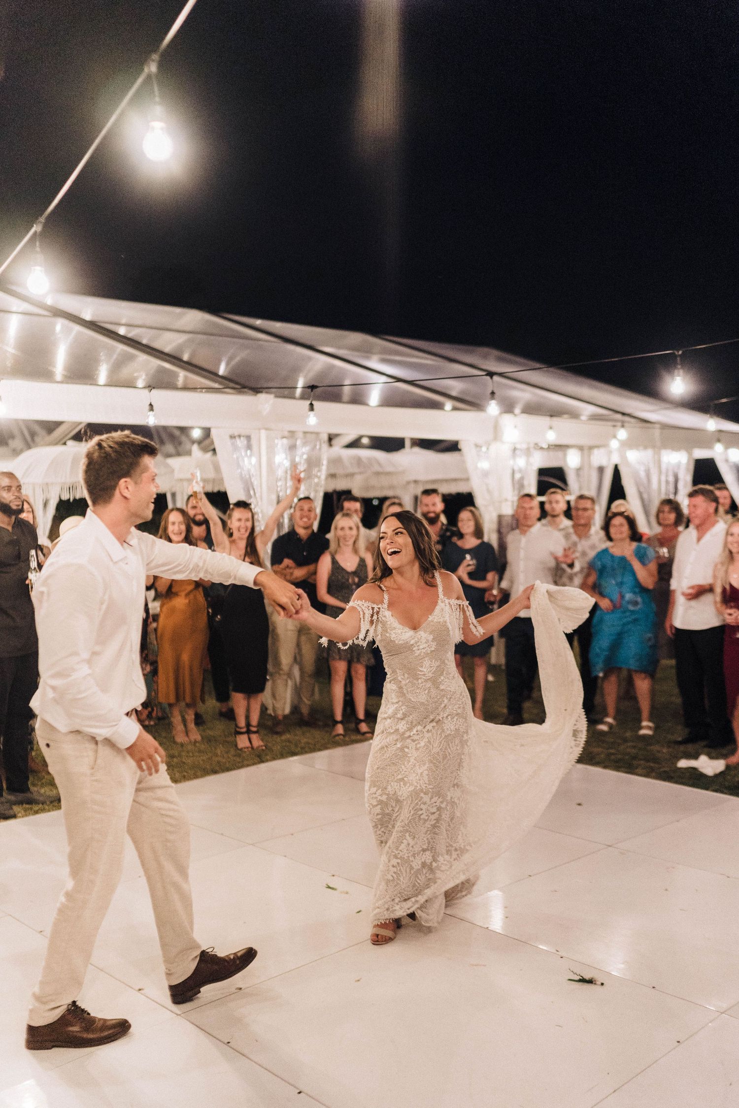 A couple shares their first dance under twinkling lights in a white tent at an evening wedding reception.