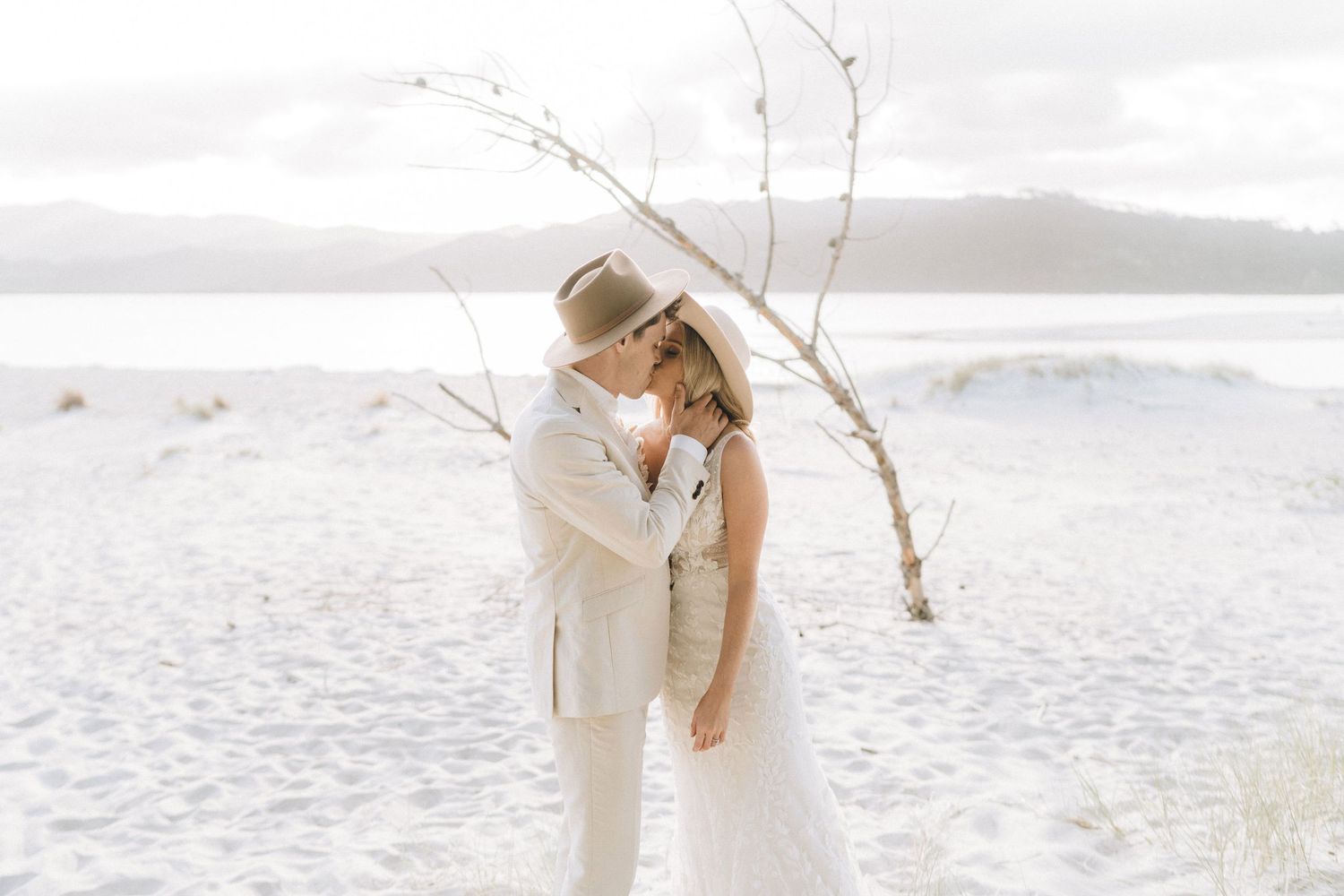 A couple in light colored wedding attire share a tender moment on a pristine white beach with mountains in the distance.
