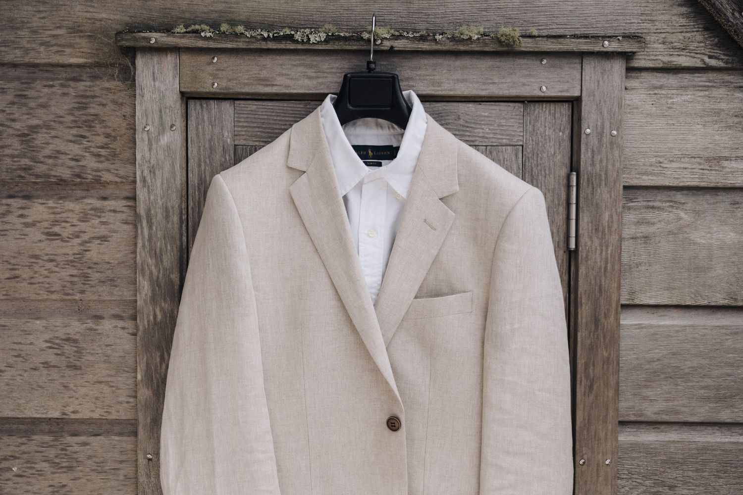 Beige linen blazer and white dress shirt hanging on wooden hanger against rustic wooden wall background.
