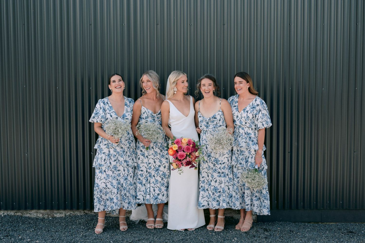 Bridesmaids in matching blue and white floral dresses pose along a green metal wall at an outdoor wedding event.