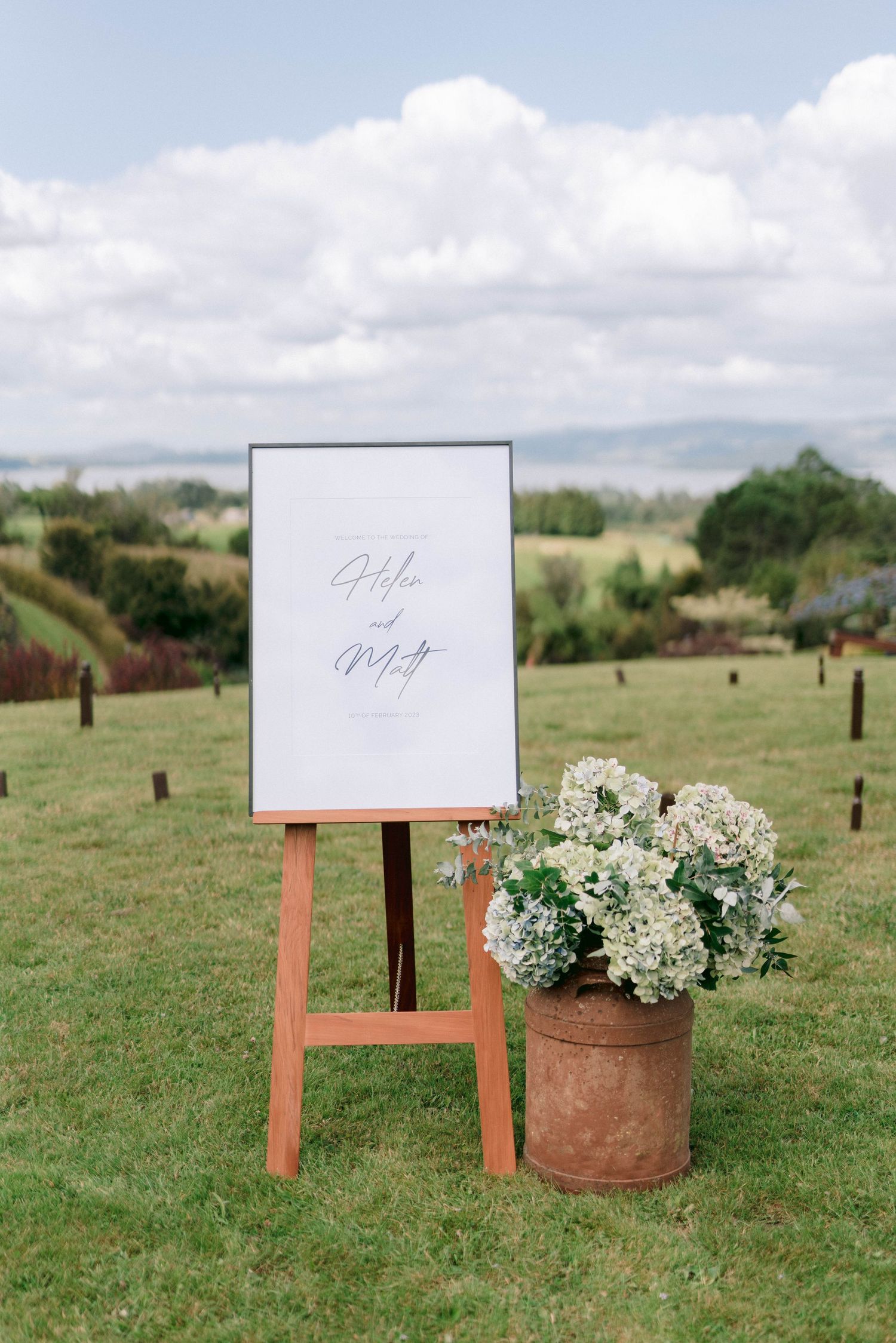 A wooden easel with a blank sign stands in a grassy outdoor setting with a potted hydrangea arrangement nearby.
