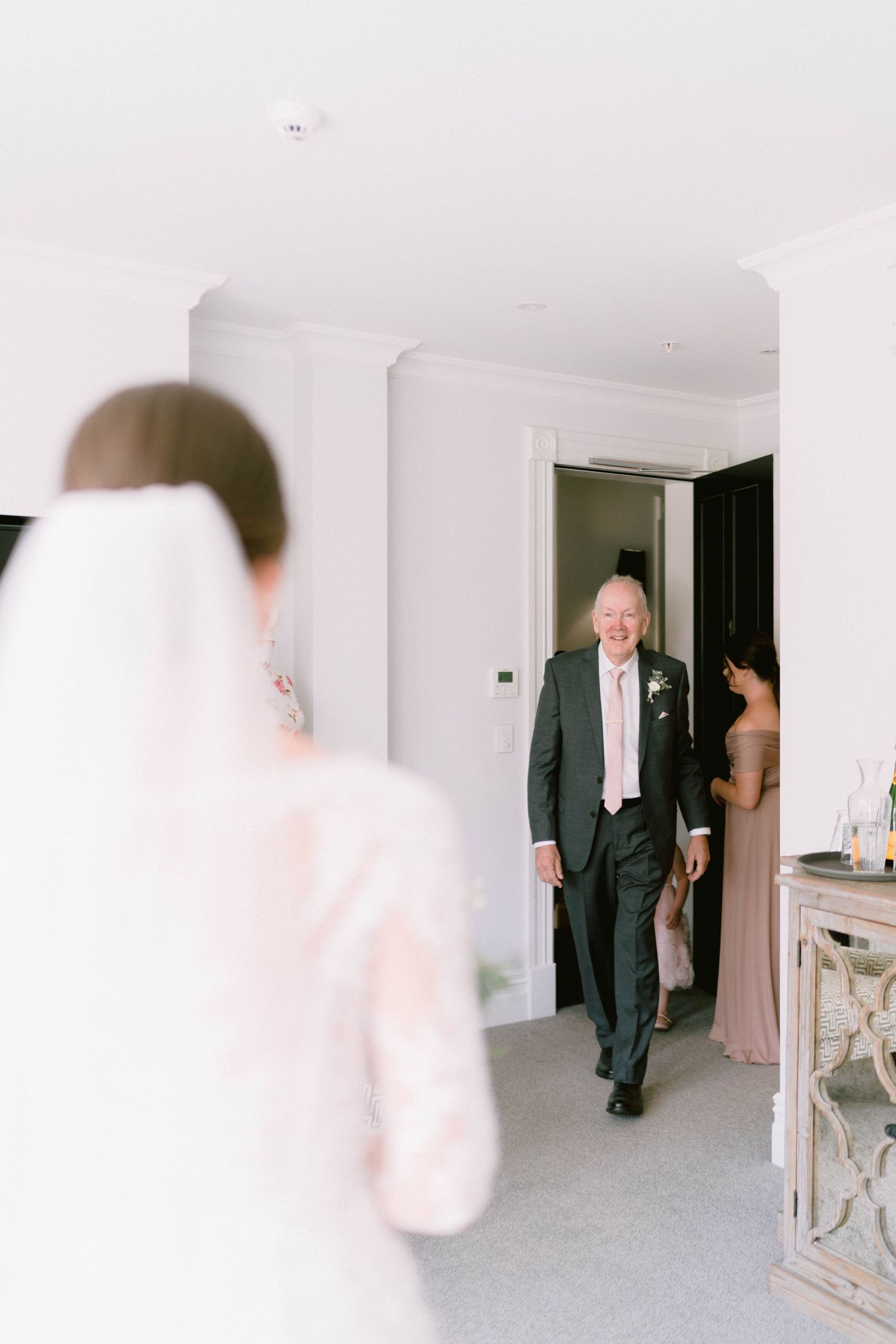 A wedding party moment captured inside a bright white room as someone enters through a doorway.