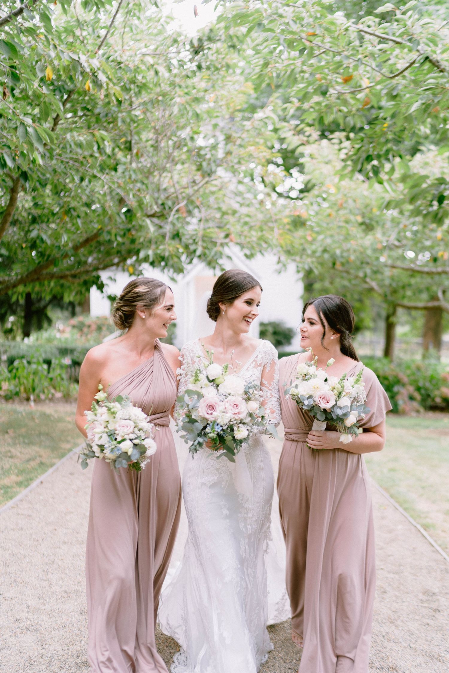Three attendants in blush pink dresses walk together on a garden path while holding white floral bouquets.
