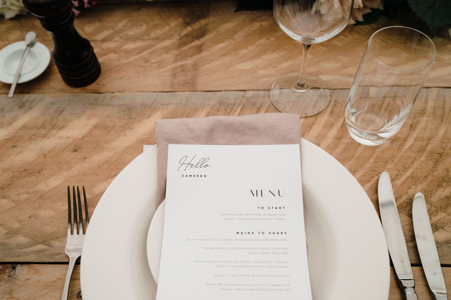 Elegant wedding menu card placed on white plate at rustic wooden table setting with silverware and glasses.