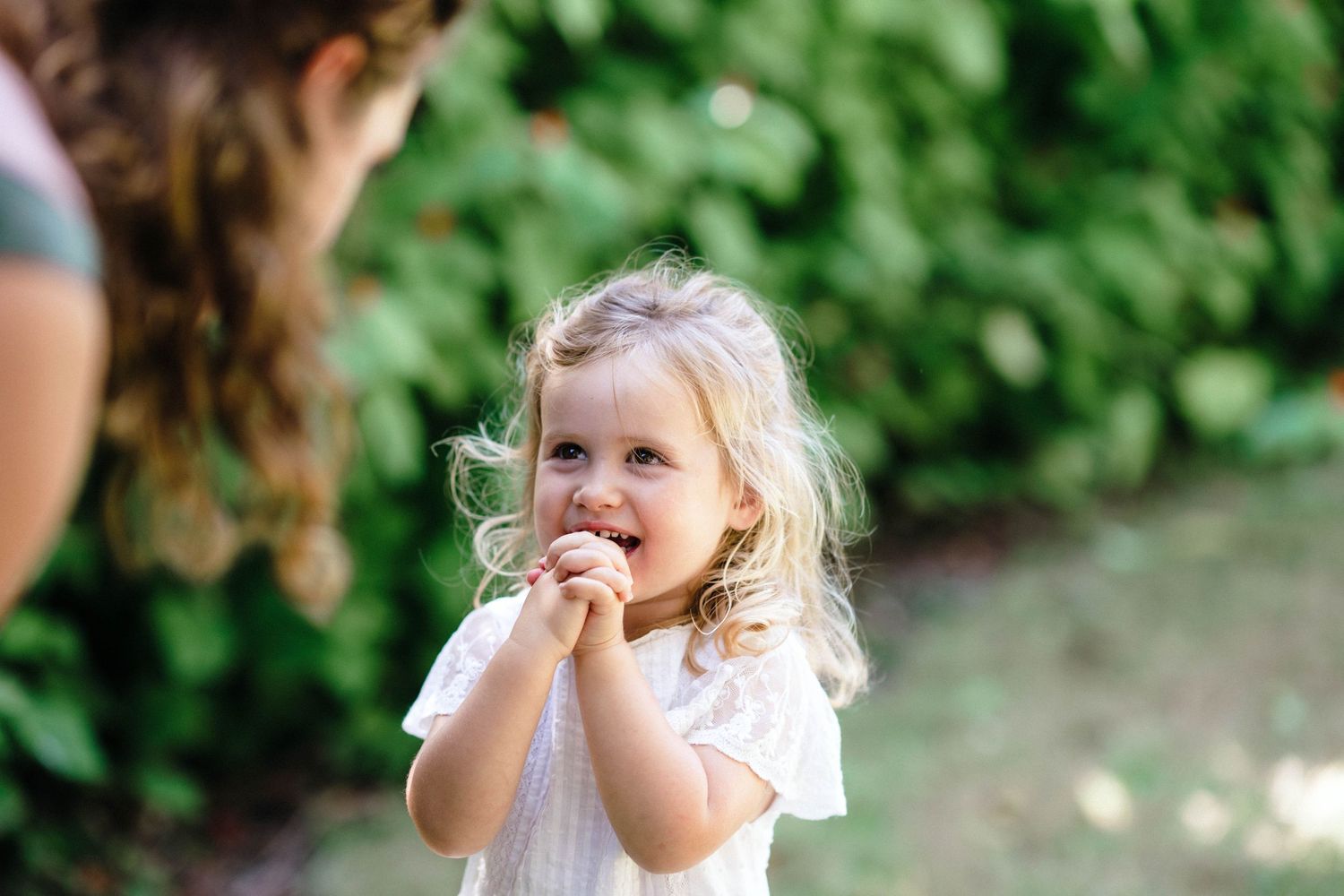 Young child in a white dress standing outdoors with a playful expression against green foliage background.