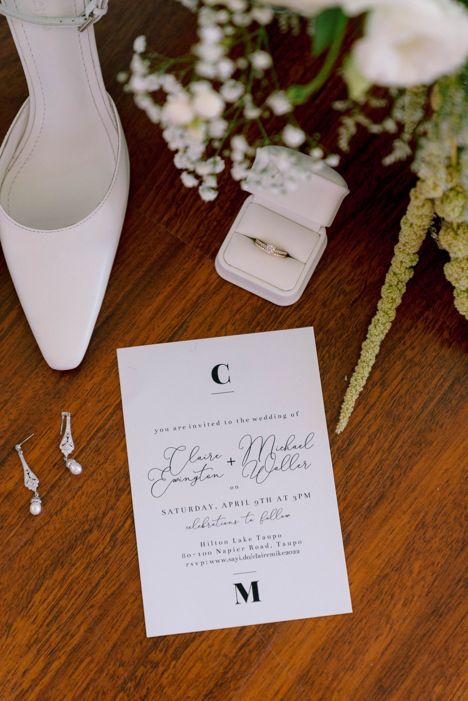 Wedding details flat lay with white invitation, ring box, bridal shoe and delicate flowers on wooden surface.