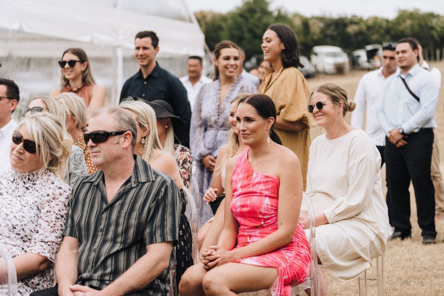 Wedding guests seated outdoors wearing sunglasses and summer attire.