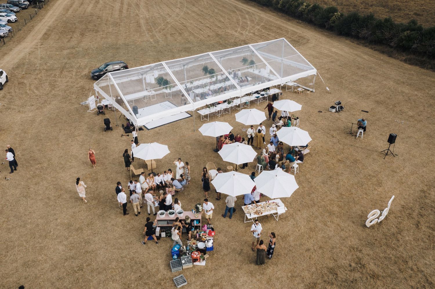 Aerial view of outdoor wedding venue with clear tent and round tables on brown field.