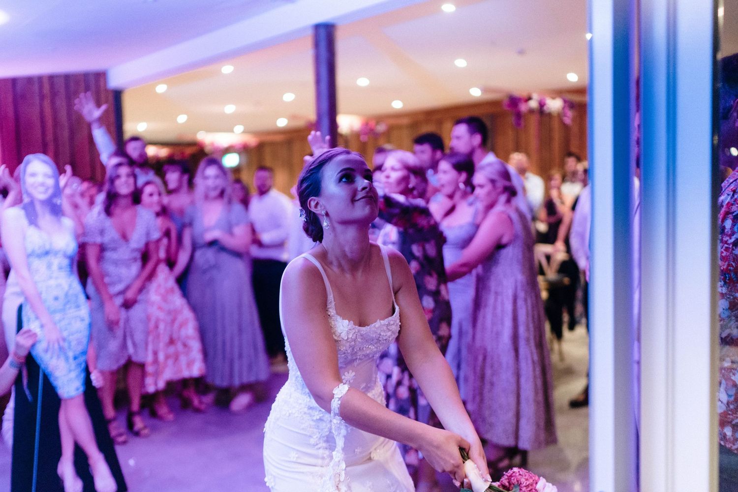 Wedding celebration on a dance floor with purple lighting as a bouquet is about to be tossed.