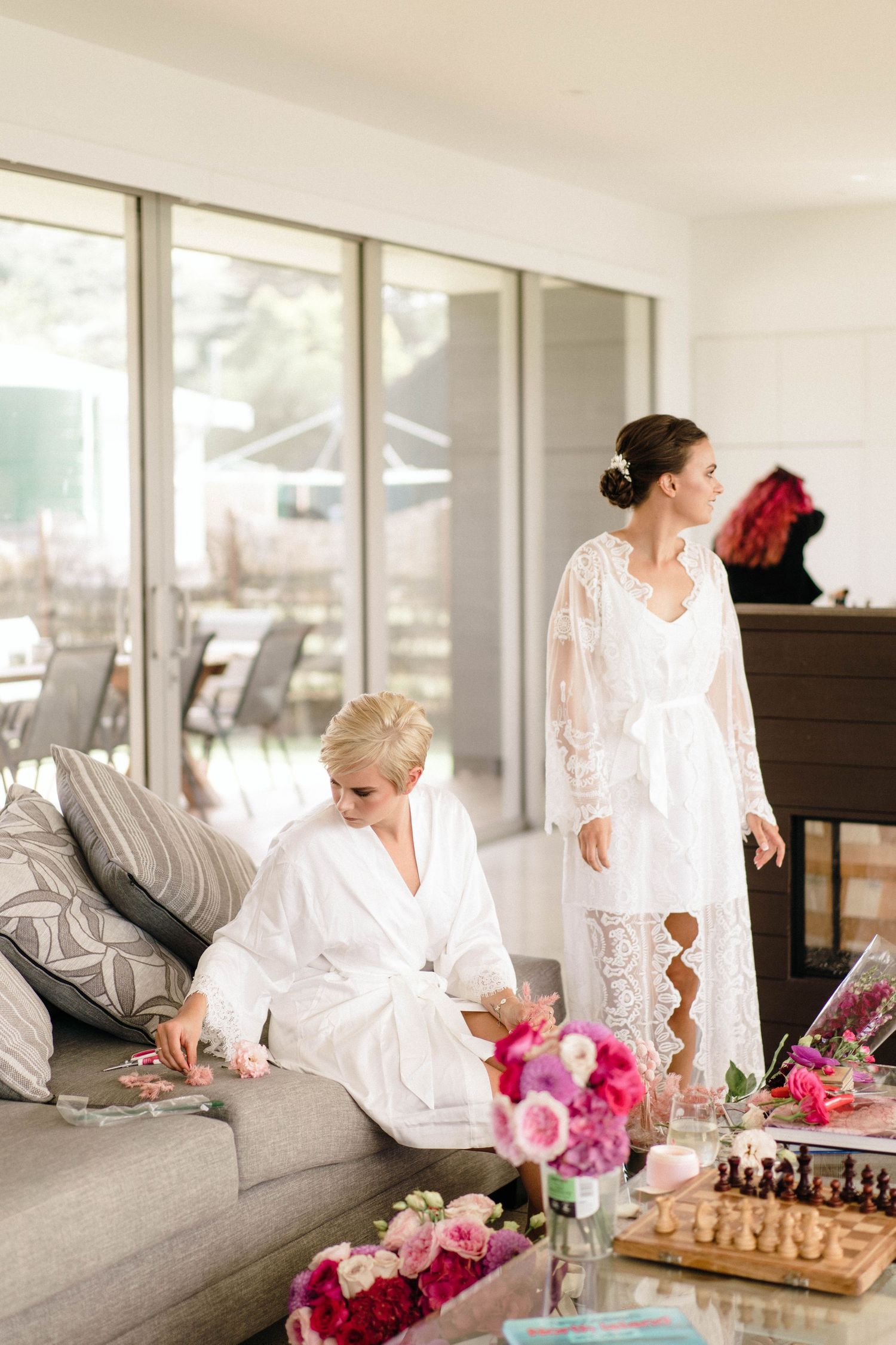 Wedding party in white robes relaxing on gray couch with pink flowers and chess set.