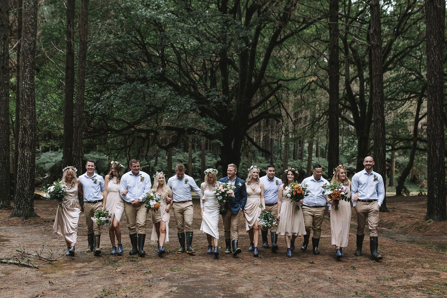 A wedding party dressed in blue and tan outfits poses along a scenic forest path in natural light.