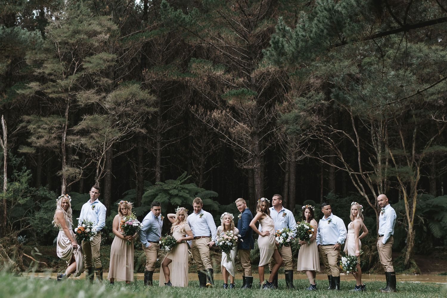 A group of wedding guests walks in a line across a wooded outdoor path while participating in wedding party photos.