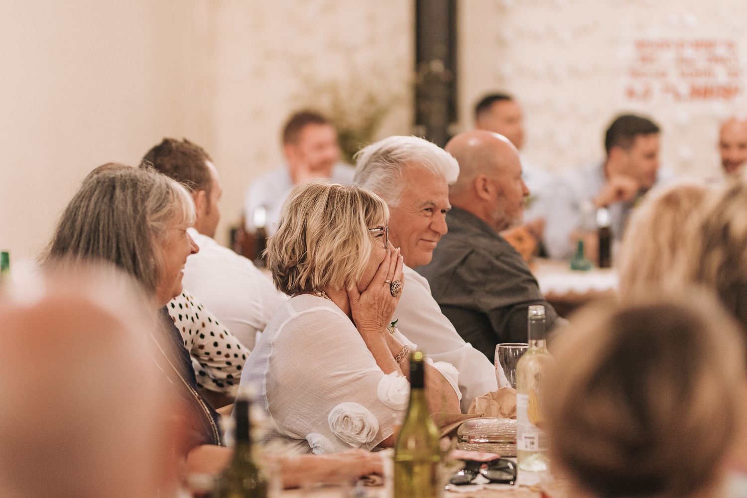 Wedding guests enjoy dinner at a softly lit reception gathering with wine bottles on tables.