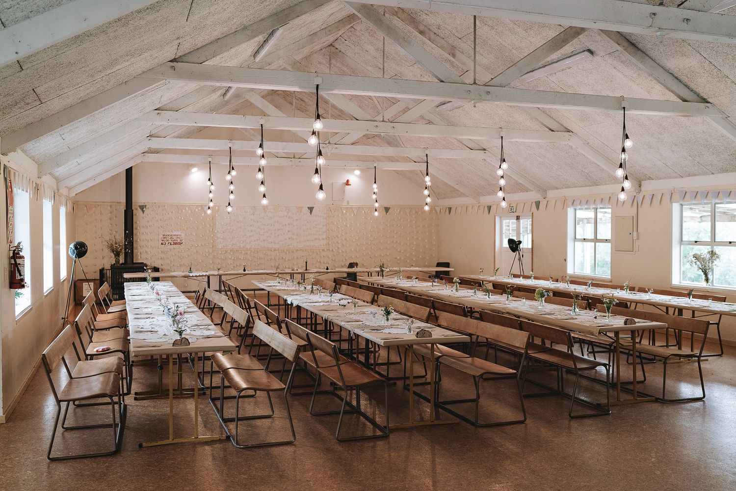 Long wooden dining tables line the interior of a bright white rustic hall with exposed wooden beams and modern lighting.