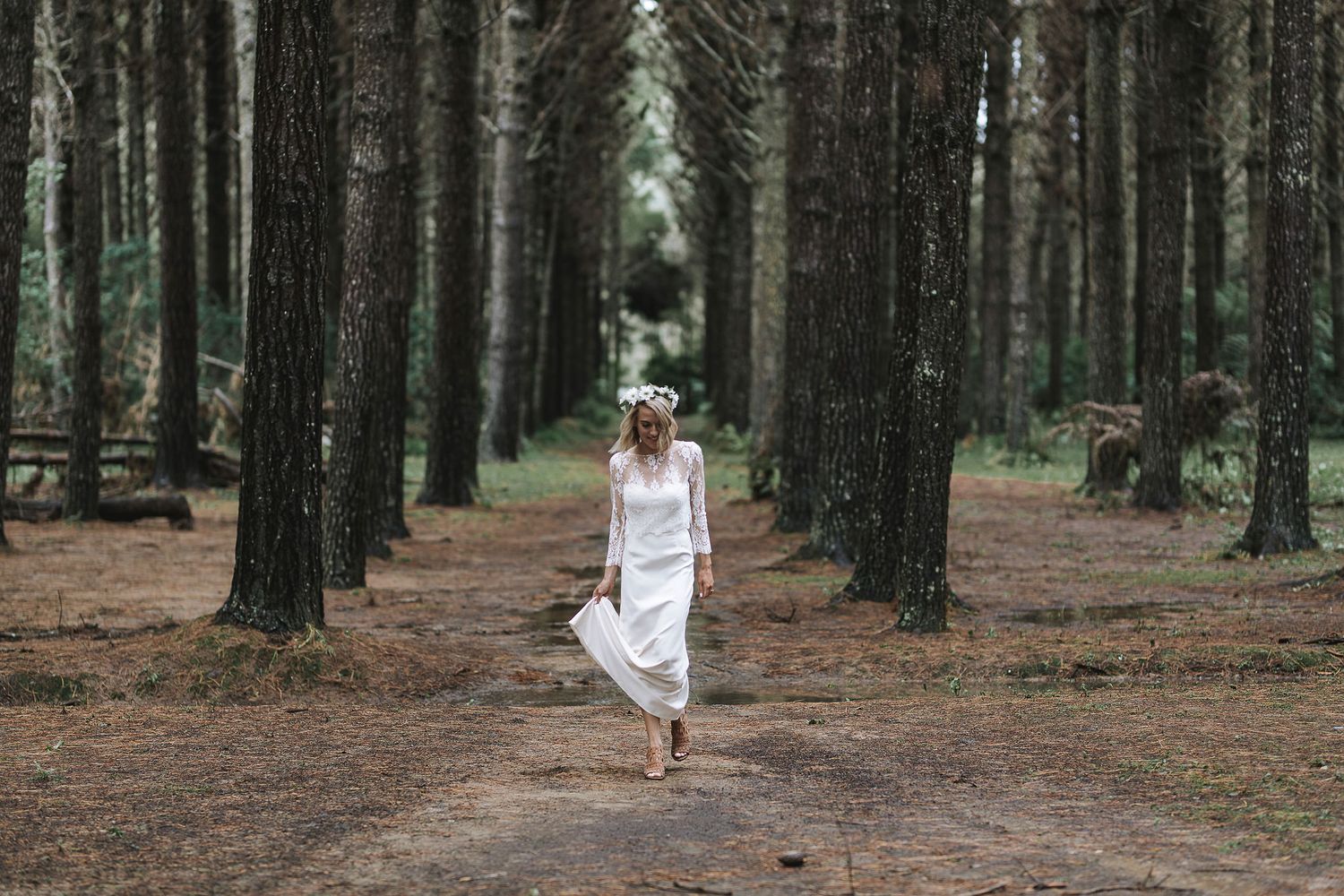 Figure in white dress walks barefoot down a tree-lined forest path between tall evergreen trees.