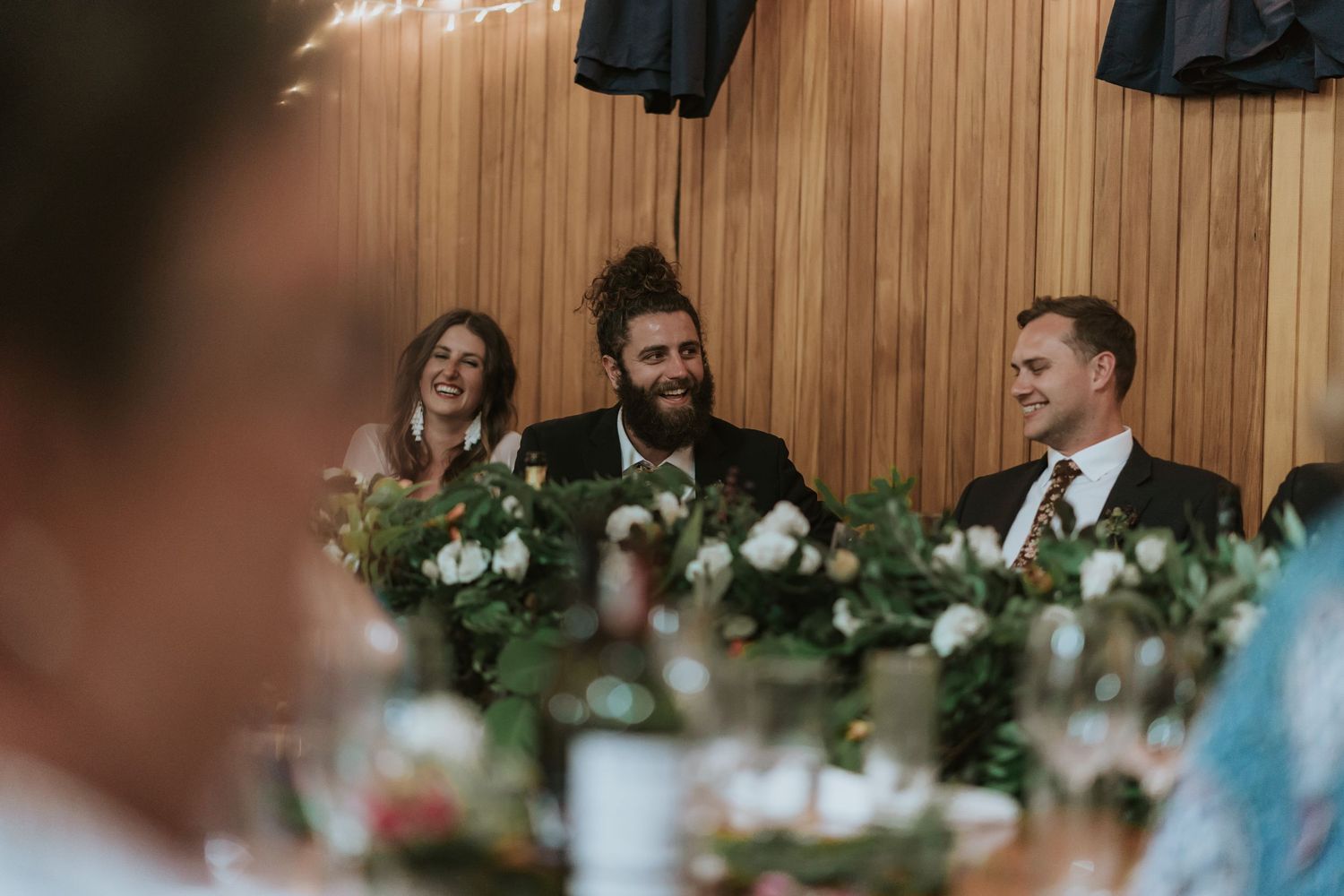 Wedding guests laugh joyfully during a reception in a rustic wood-paneled venue.