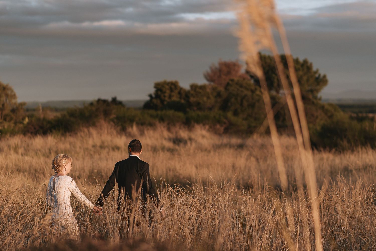Two people in wedding attire stand together in tall golden grass during sunset in a rustic outdoor setting.