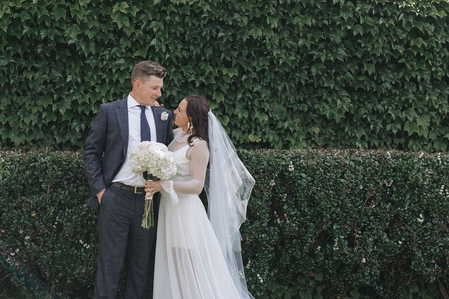A couple shares an intimate moment in front of a lush green hedge while wearing traditional wedding attire.