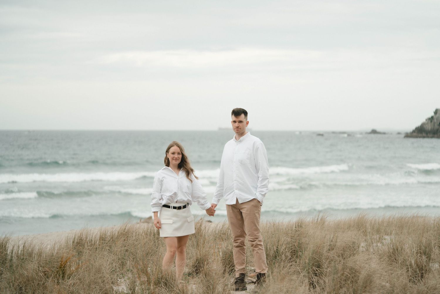 A couple stands holding hands in tall beach grass along a cloudy coastline.