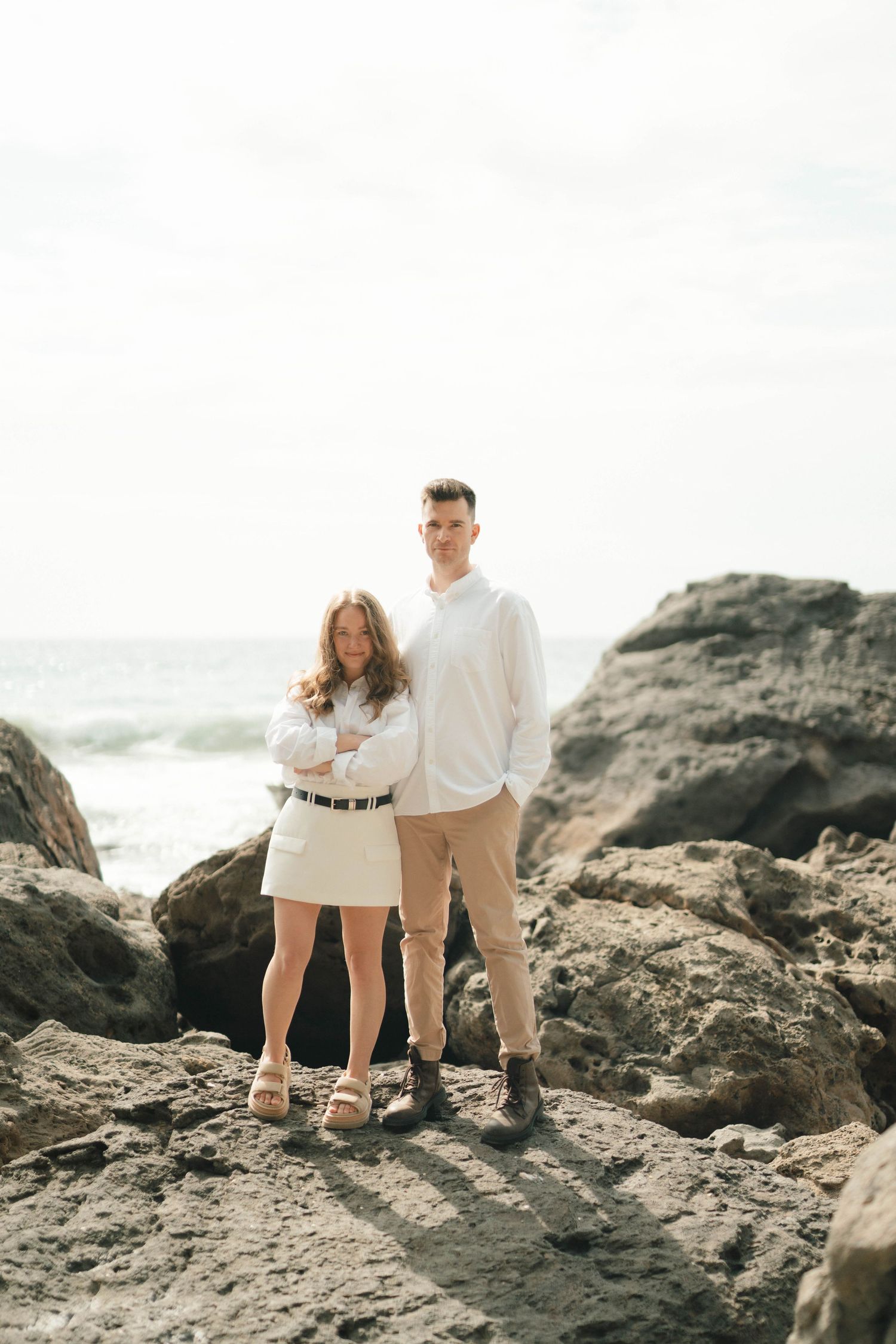 Couple in coordinated white outfits walking along rocky coastal terrain at sunset.
