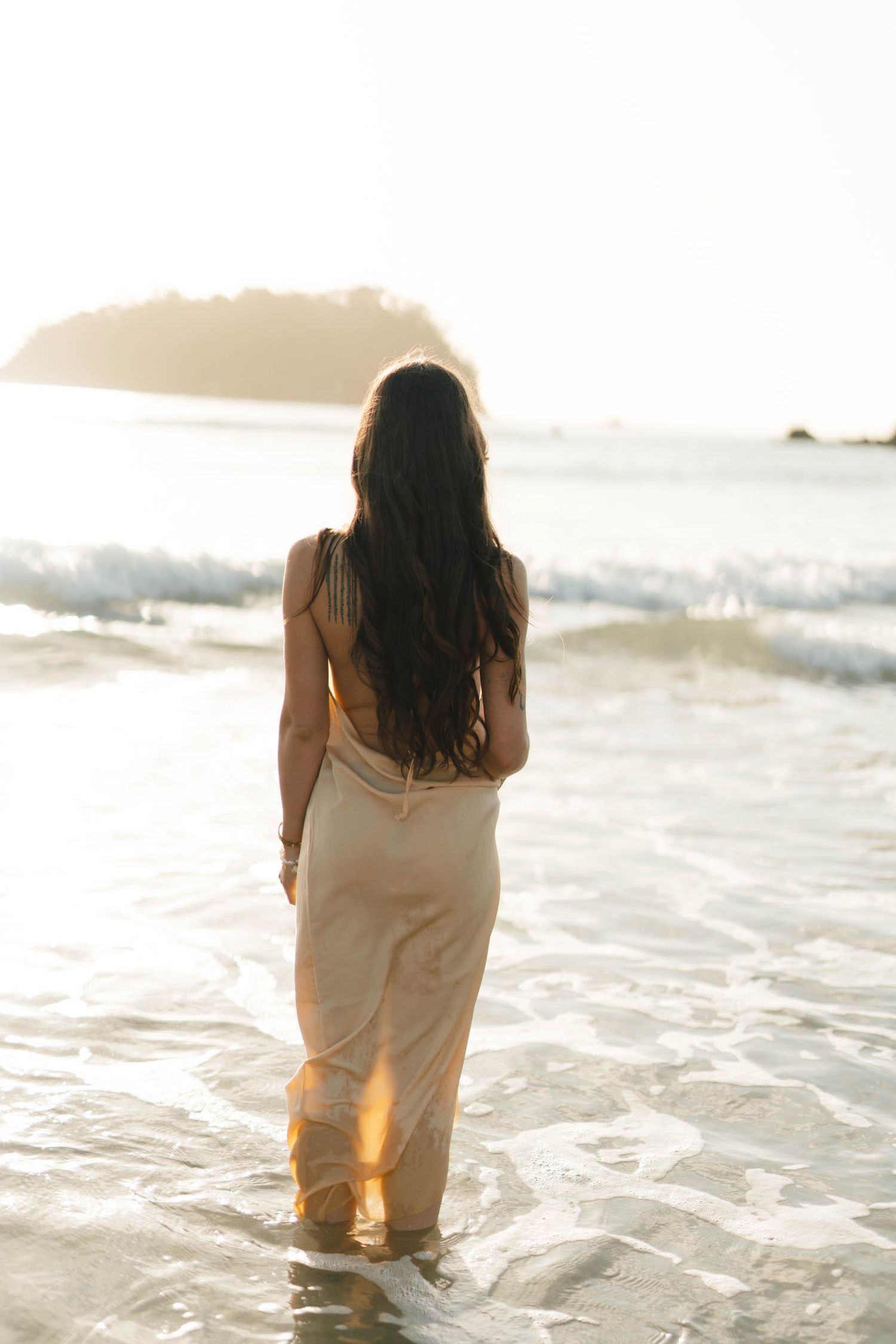 A woman in a flowing dress stands in shallow ocean waves during golden hour.