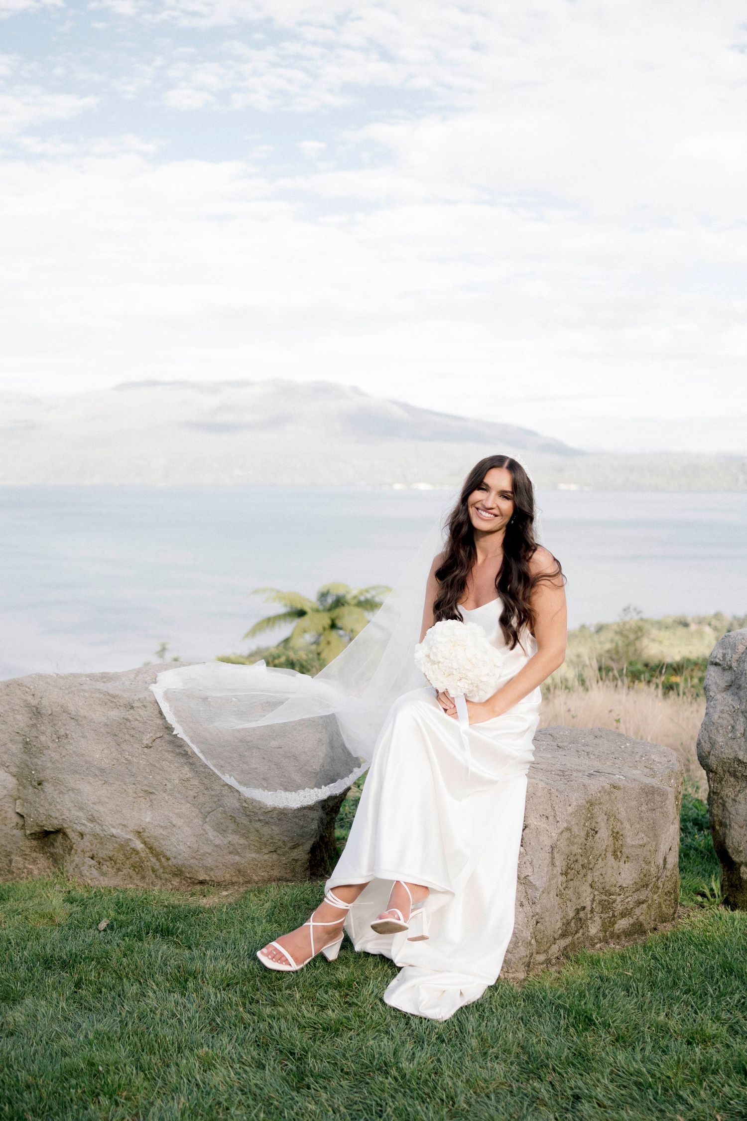 Bride in white dress poses on rocky cliff overlooking mountain lake view.