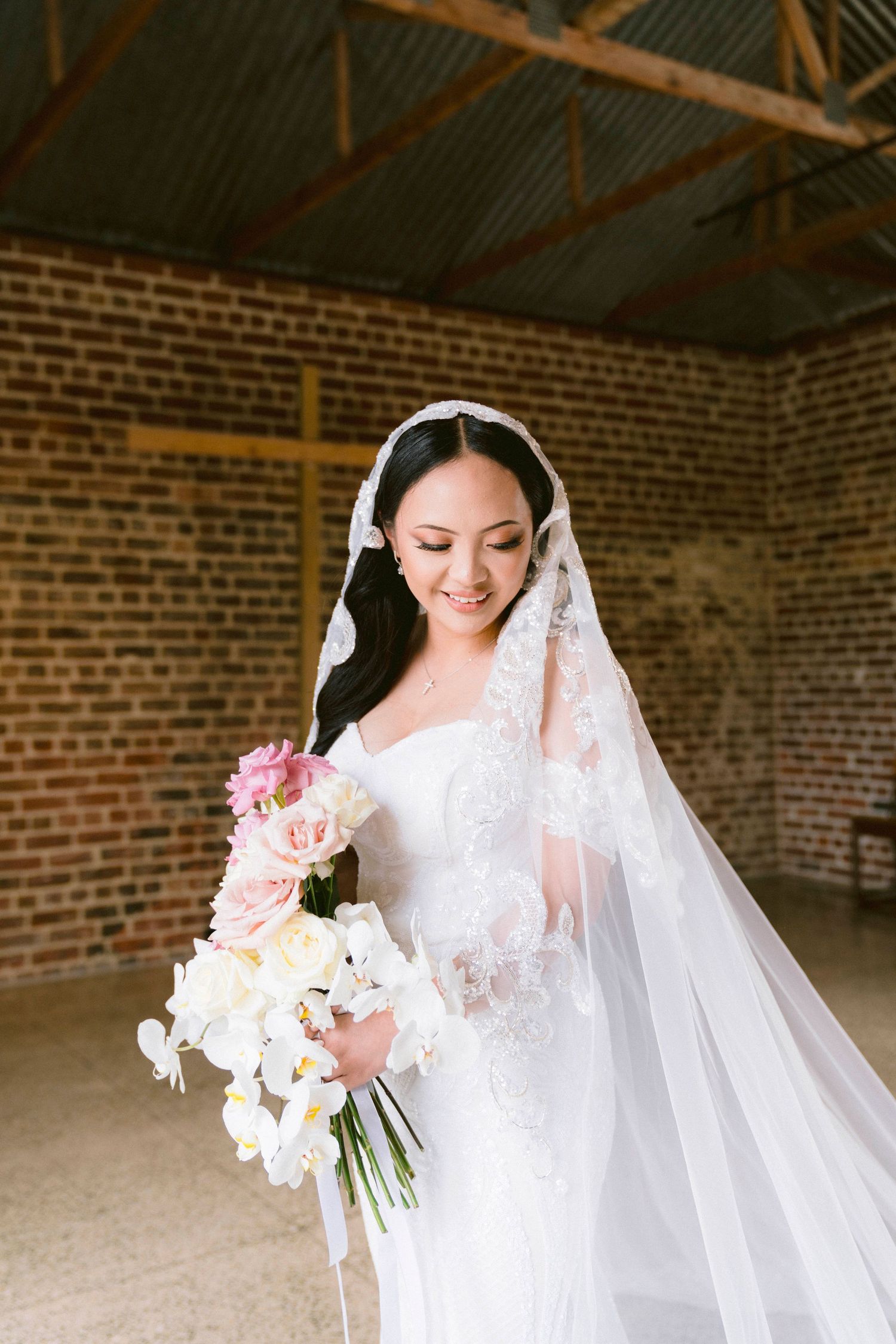 A radiant bride smiles while holding a bouquet of pink and white flowers in a vintage brick building.