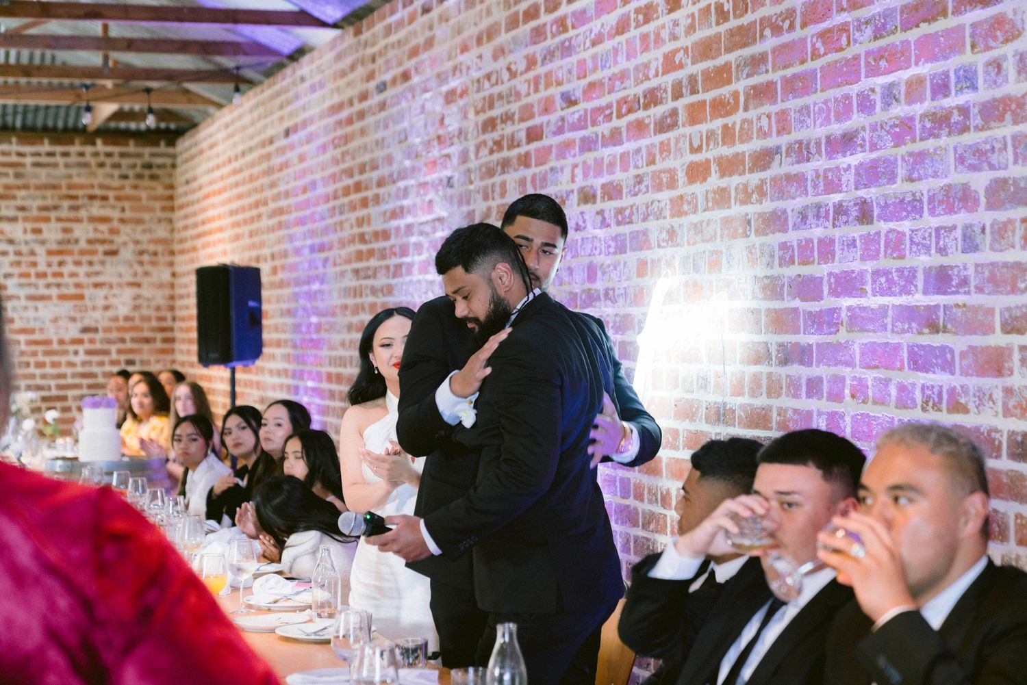 Wedding reception guests seated at long tables in a rustic brick venue with purple lighting.