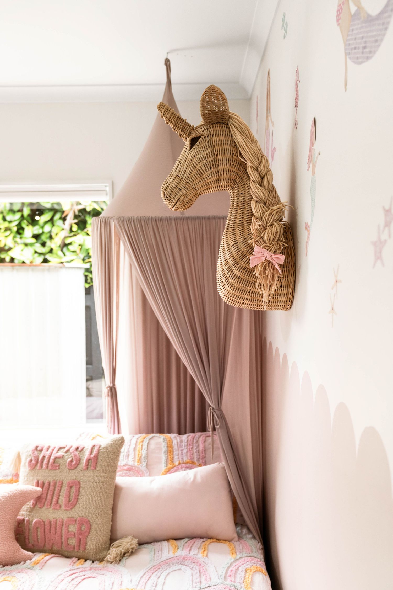 Woven rattan unicorn head wall decor with pink bow against blush curtains.